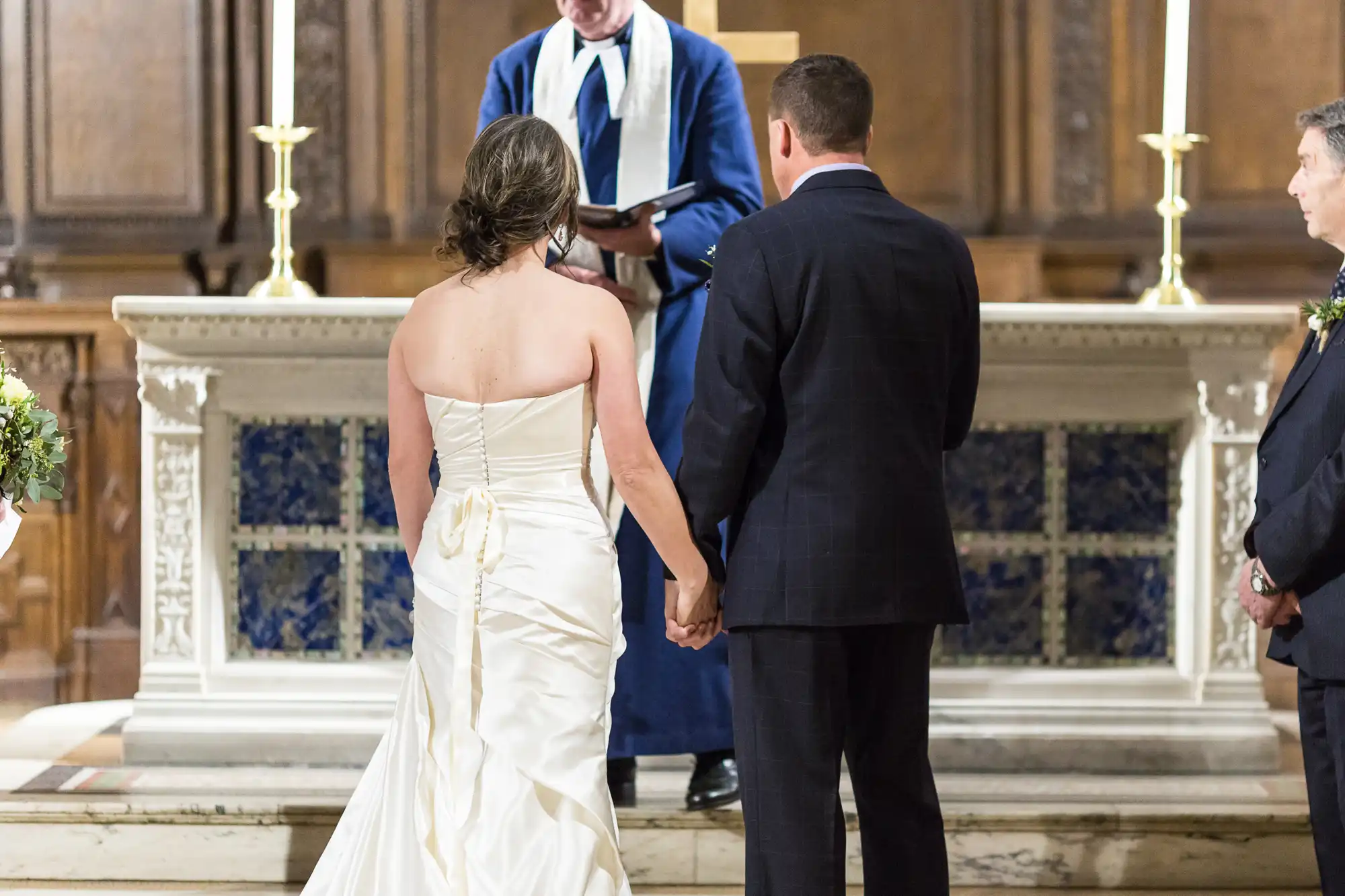 A bride and groom holding hands during a wedding ceremony in a church, with the officiant in front of them and floral decorations on the sides.