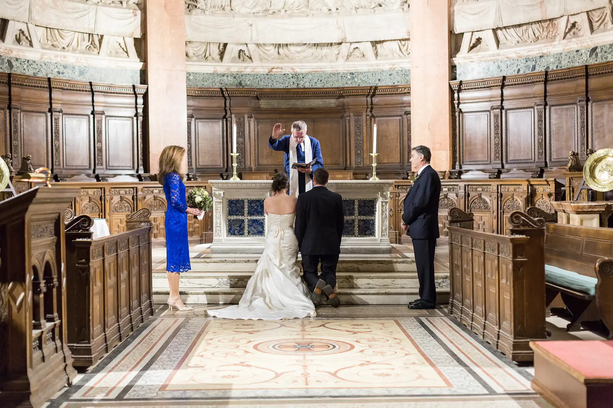 A wedding ceremony in a richly decorated church, with the bride and groom facing the officiant, and two guests watching.