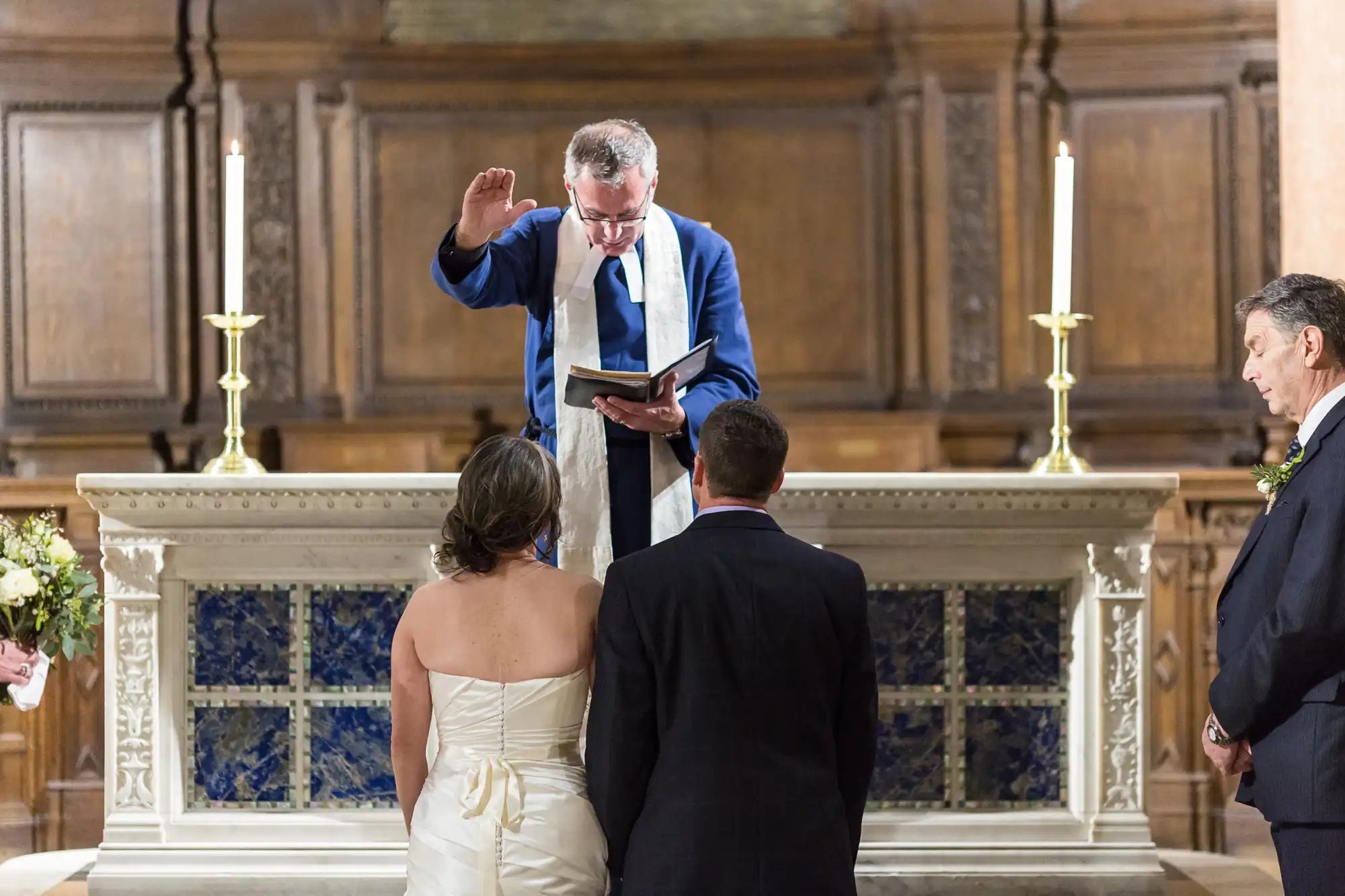 A priest conducting a wedding ceremony for a couple inside a church, with lit candles on an altar.
