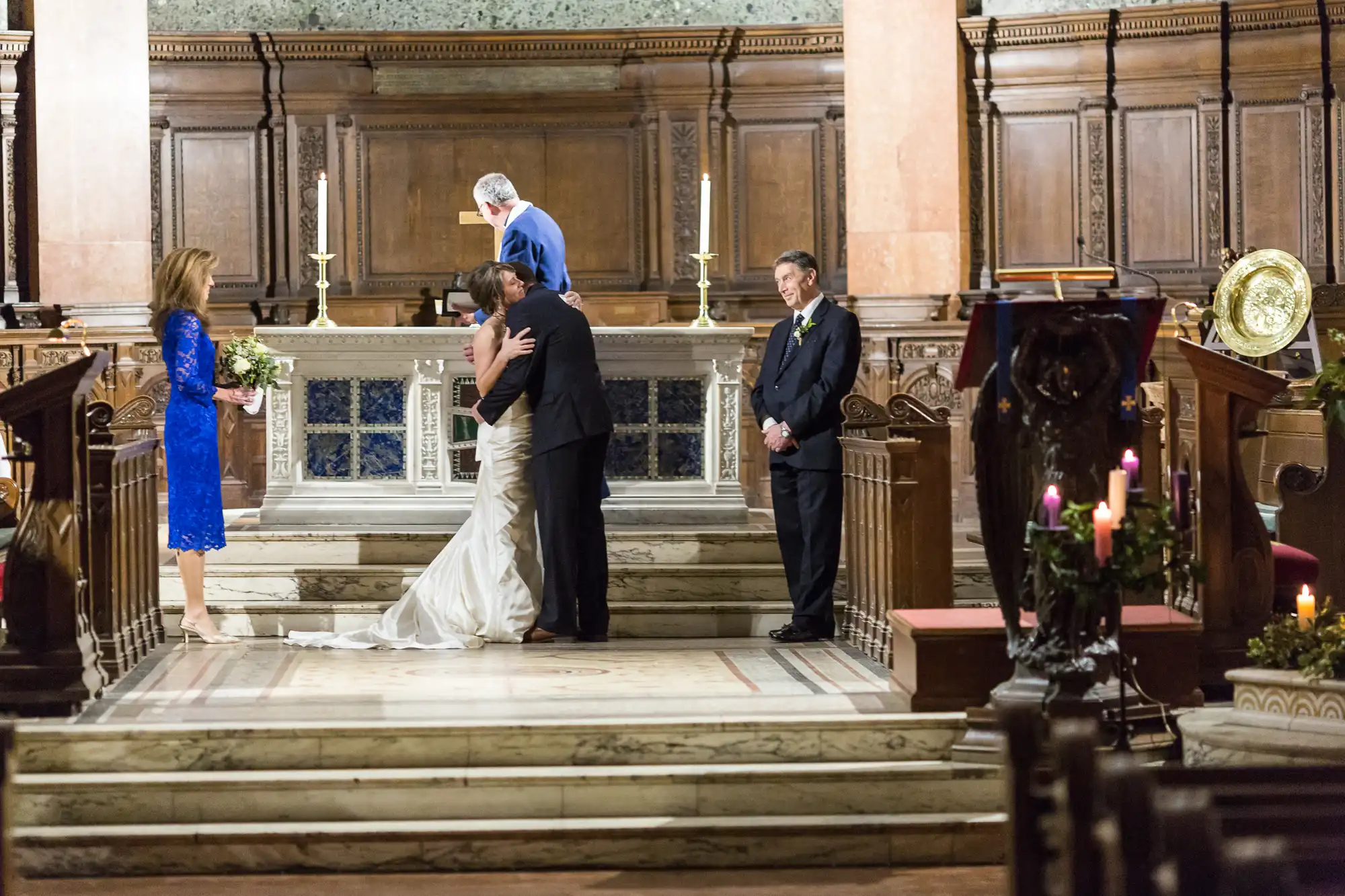 A bride and groom embracing at the altar in a church, with an officiant and two guests watching.