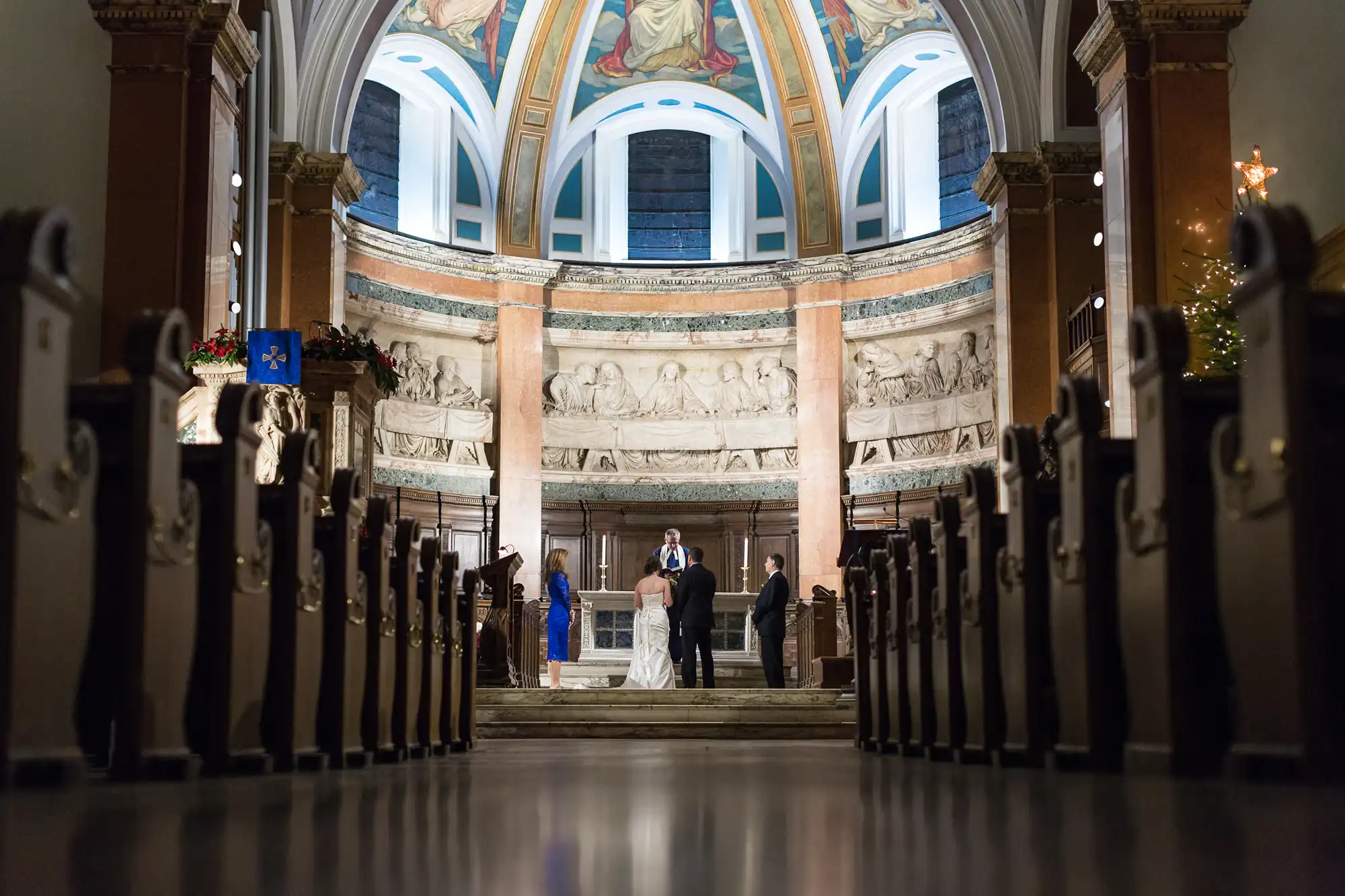 A wedding ceremony inside a church with guests seated along the aisle leading to a couple standing before an officiant under a detailed, arched altar.