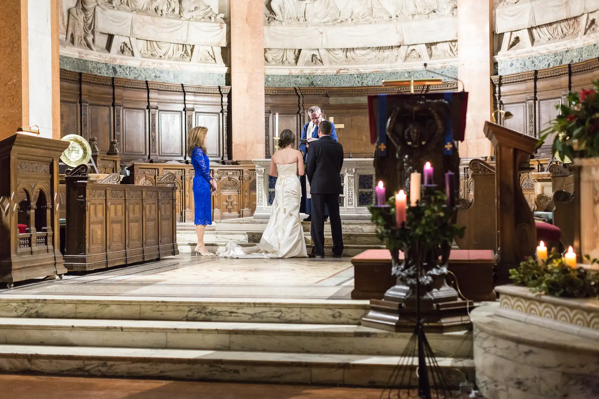 A wedding ceremony in a grand church with a couple standing before an officiant, flanked by two guests, surrounded by candles and marble architecture.