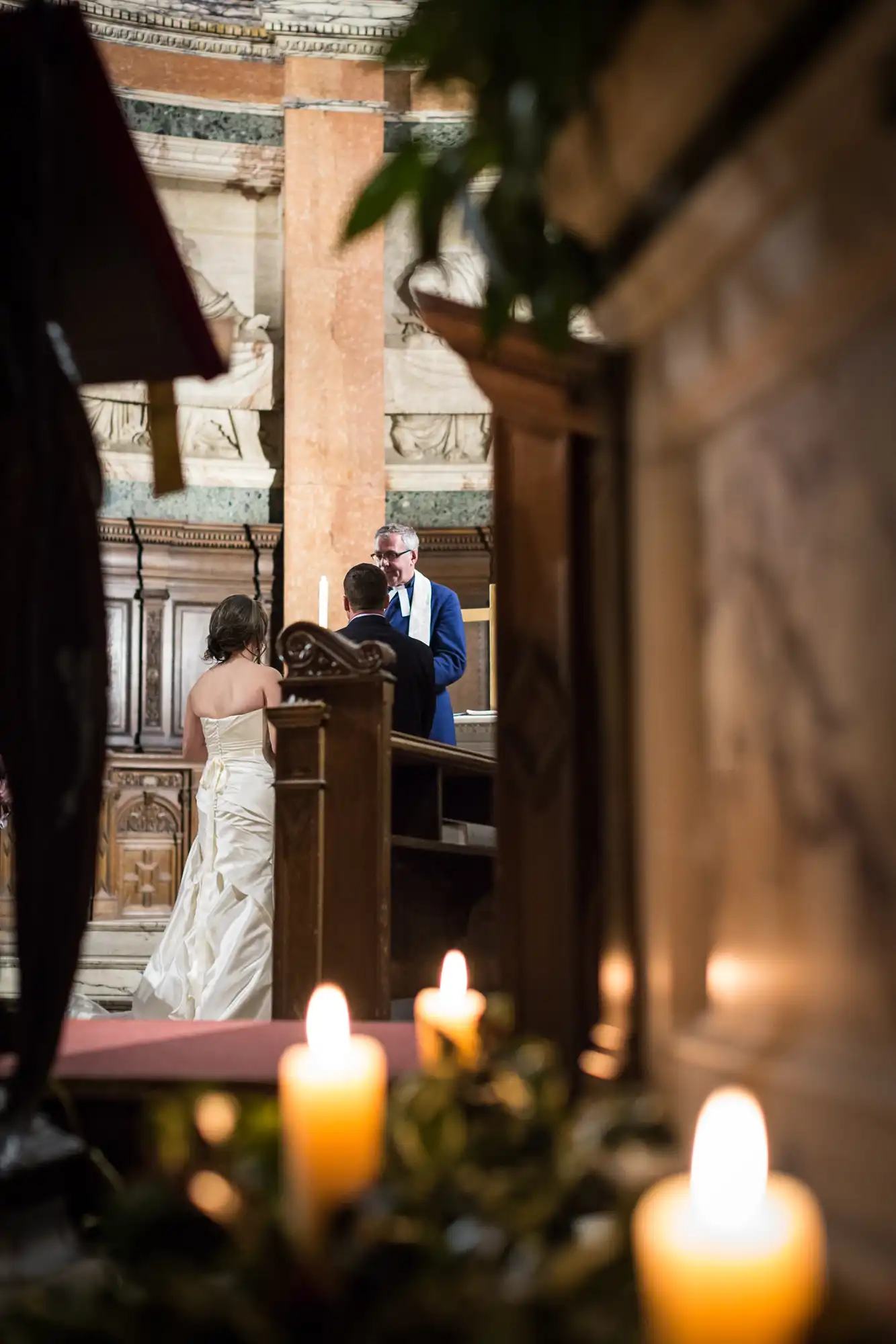 A bride and groom exchange vows at an altar, viewed through a gap between candles, in an ornate church setting.