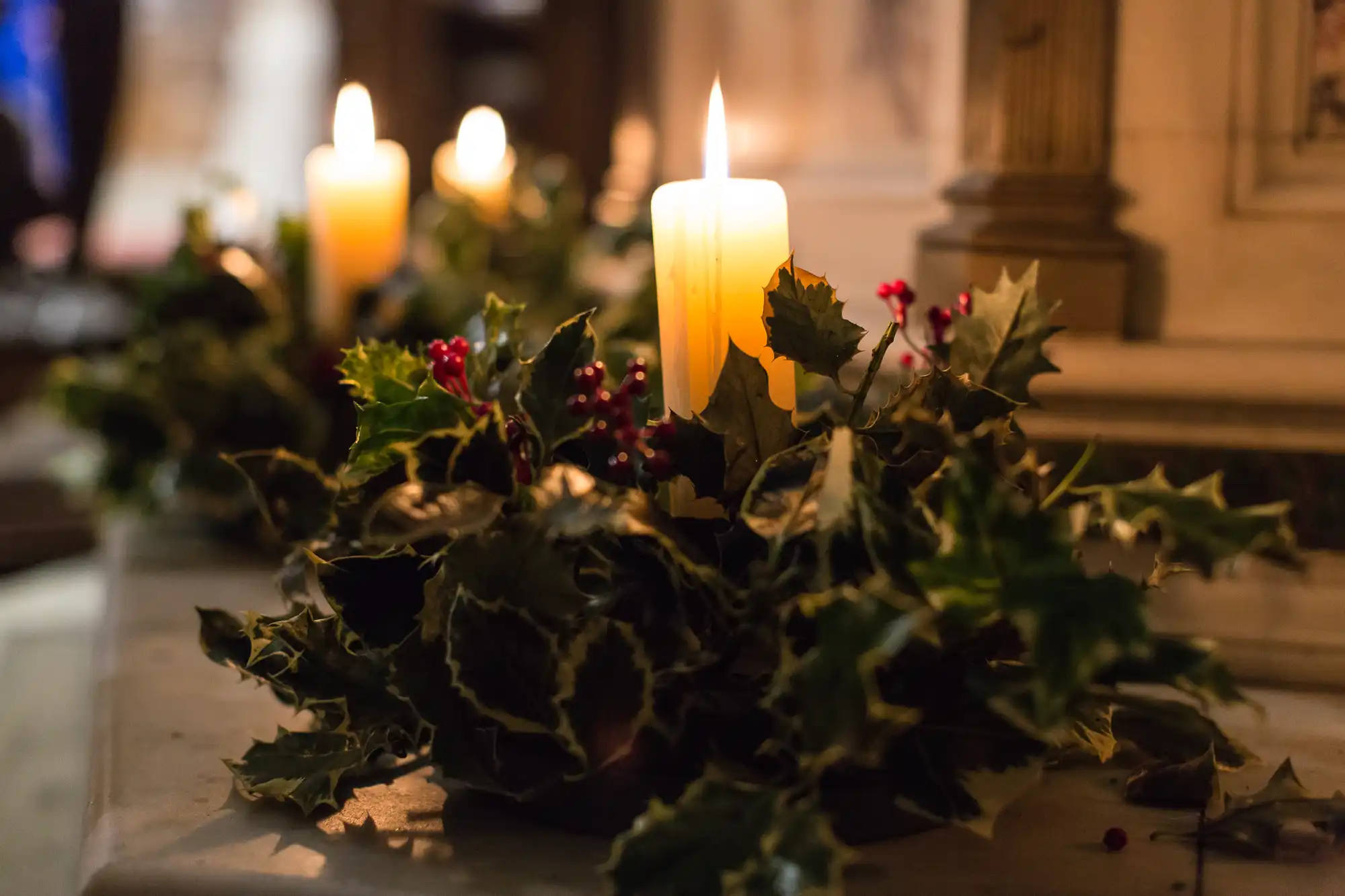 Lit candles surrounded by holly and ivy on a church altar, casting a warm glow in a dimly lit setting.