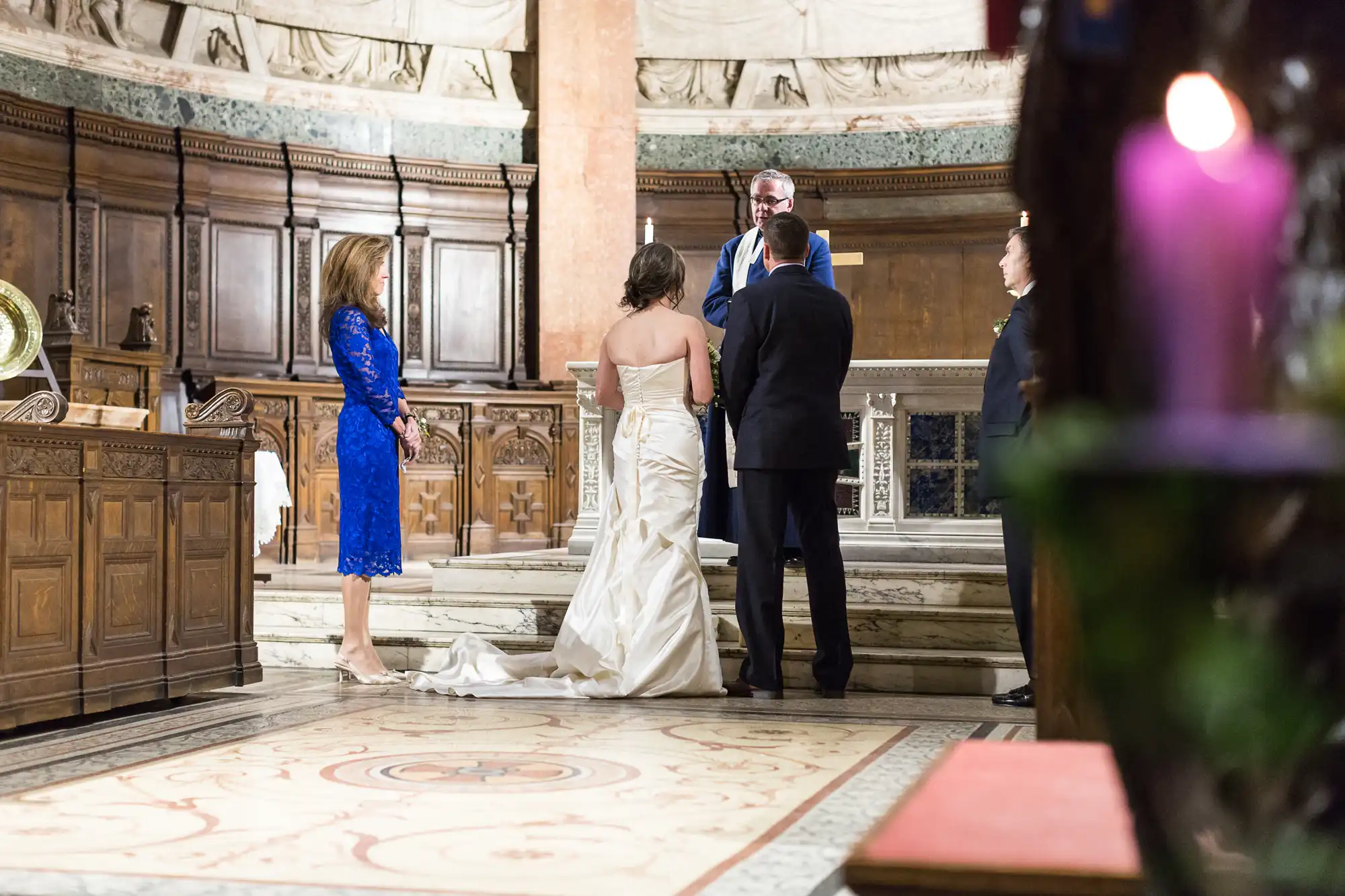 A wedding ceremony in a grand, ornate interior with a couple facing an officiant, flanked by a woman in a blue dress.