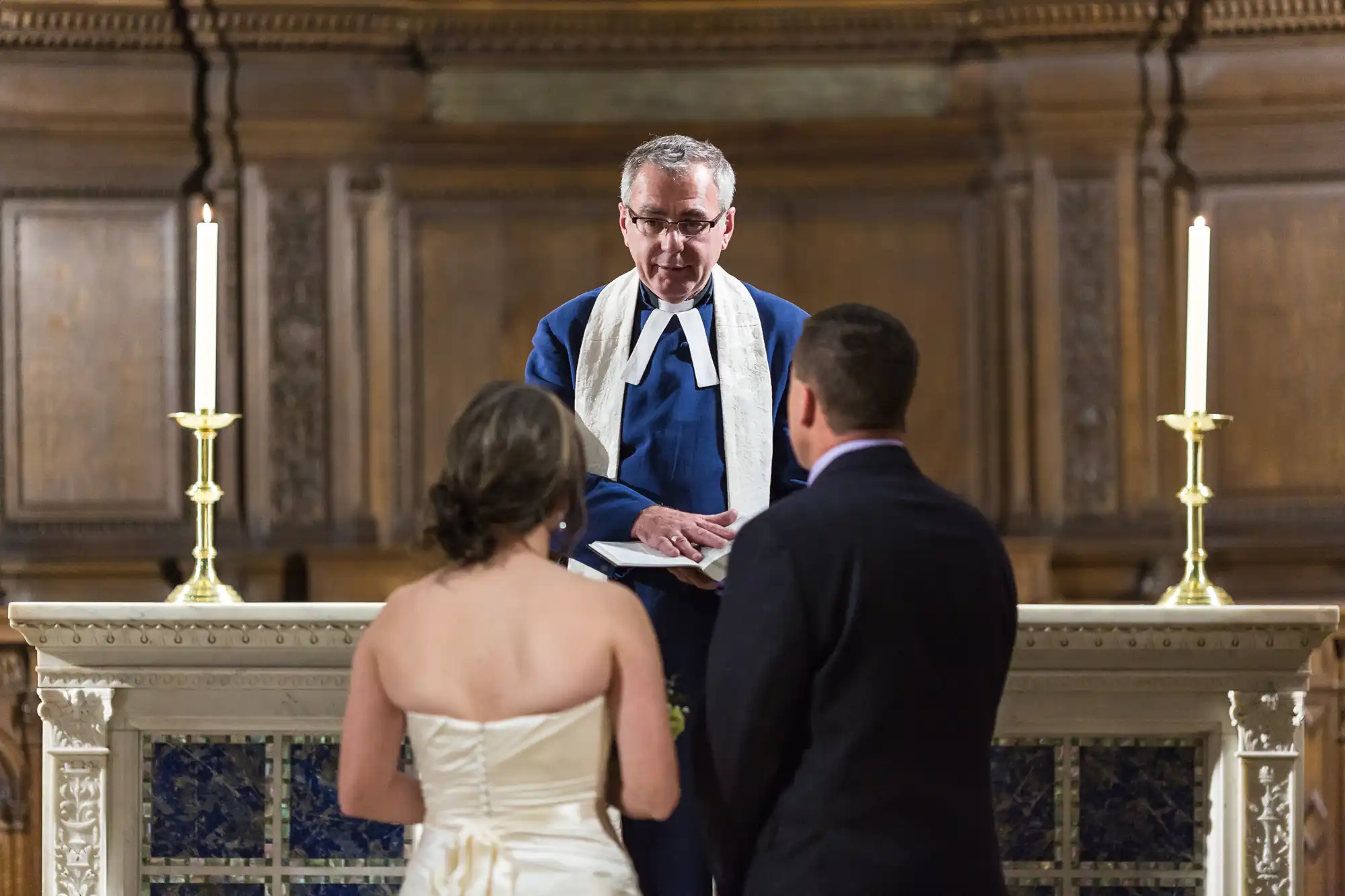 A minister officiates a wedding ceremony for a couple in a church, standing behind an altar with candles.