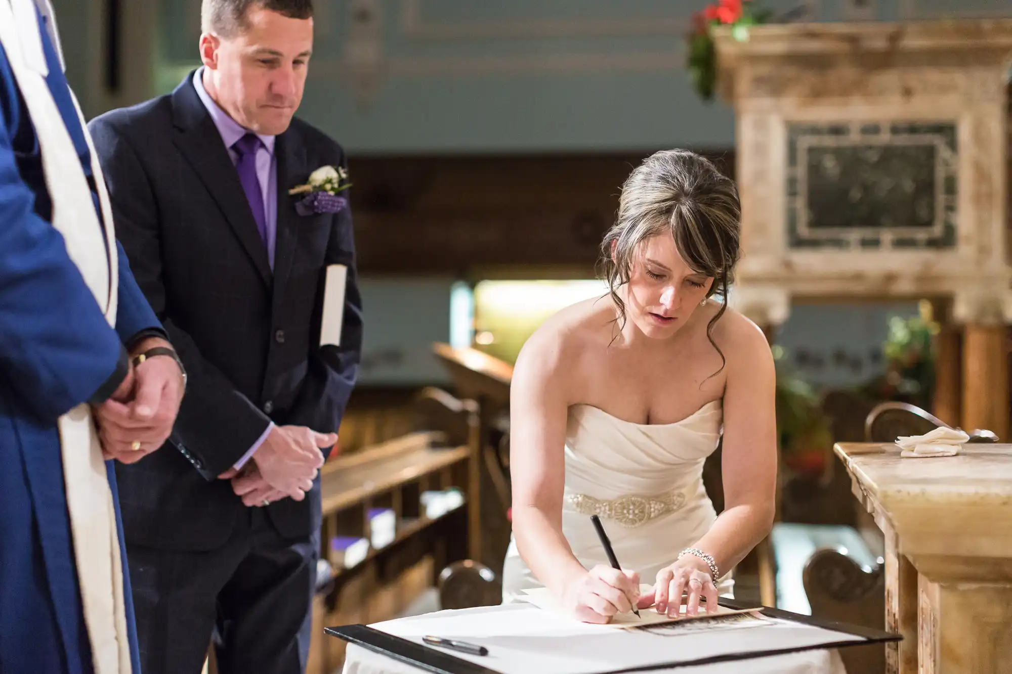 A bride in a strapless dress signs a document at her wedding ceremony, with an onlooking man in a suit and purple boutonniere.