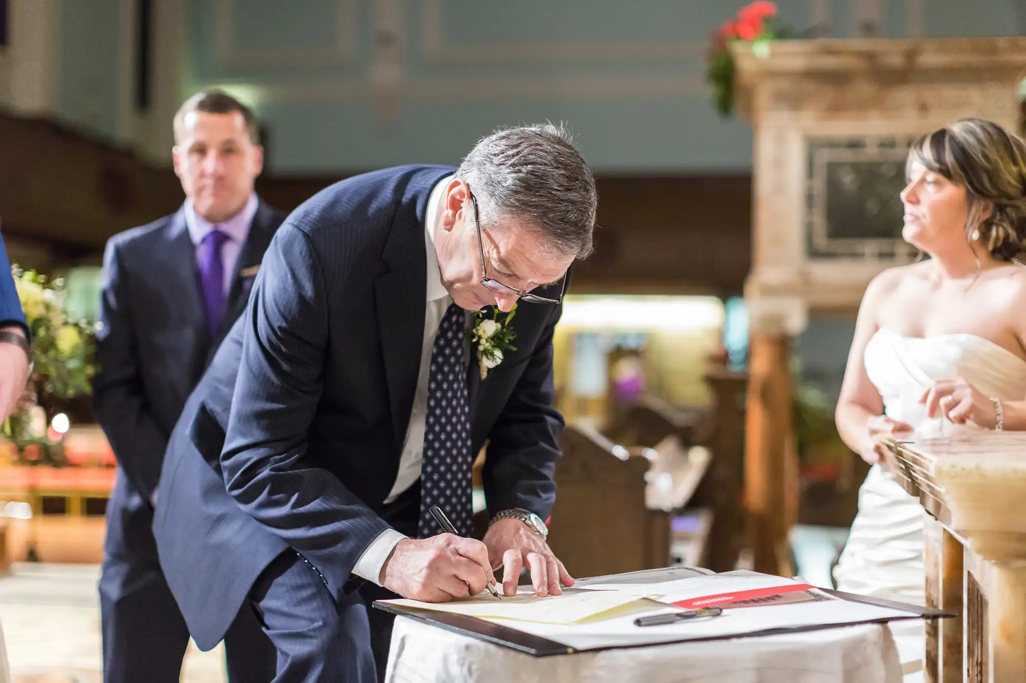 An older man signs a document at a wedding ceremony, watched by a man in a suit and a woman in a wedding dress.