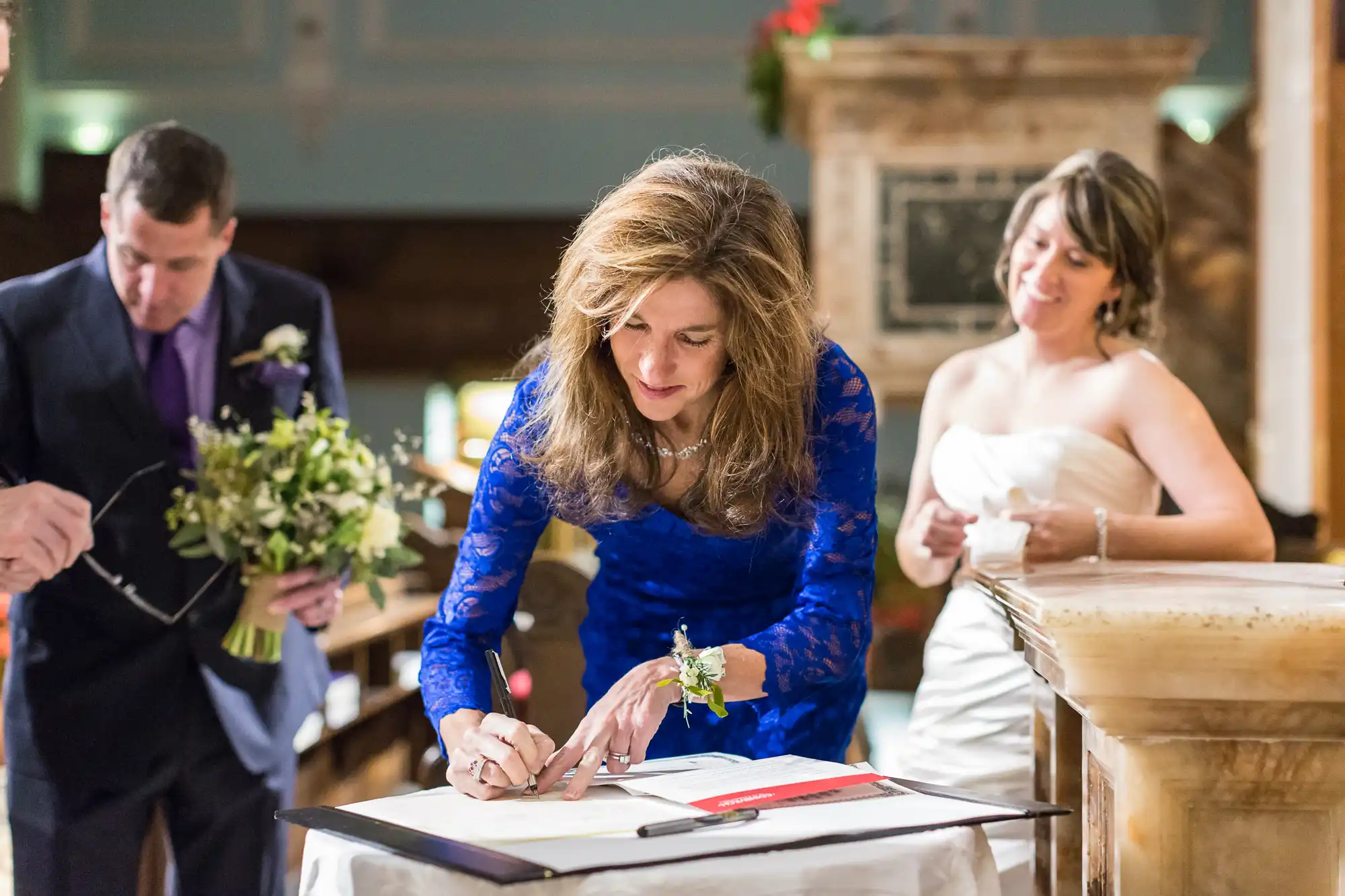 A woman in a blue dress signs a document at a wedding ceremony with a couple and another man standing nearby.