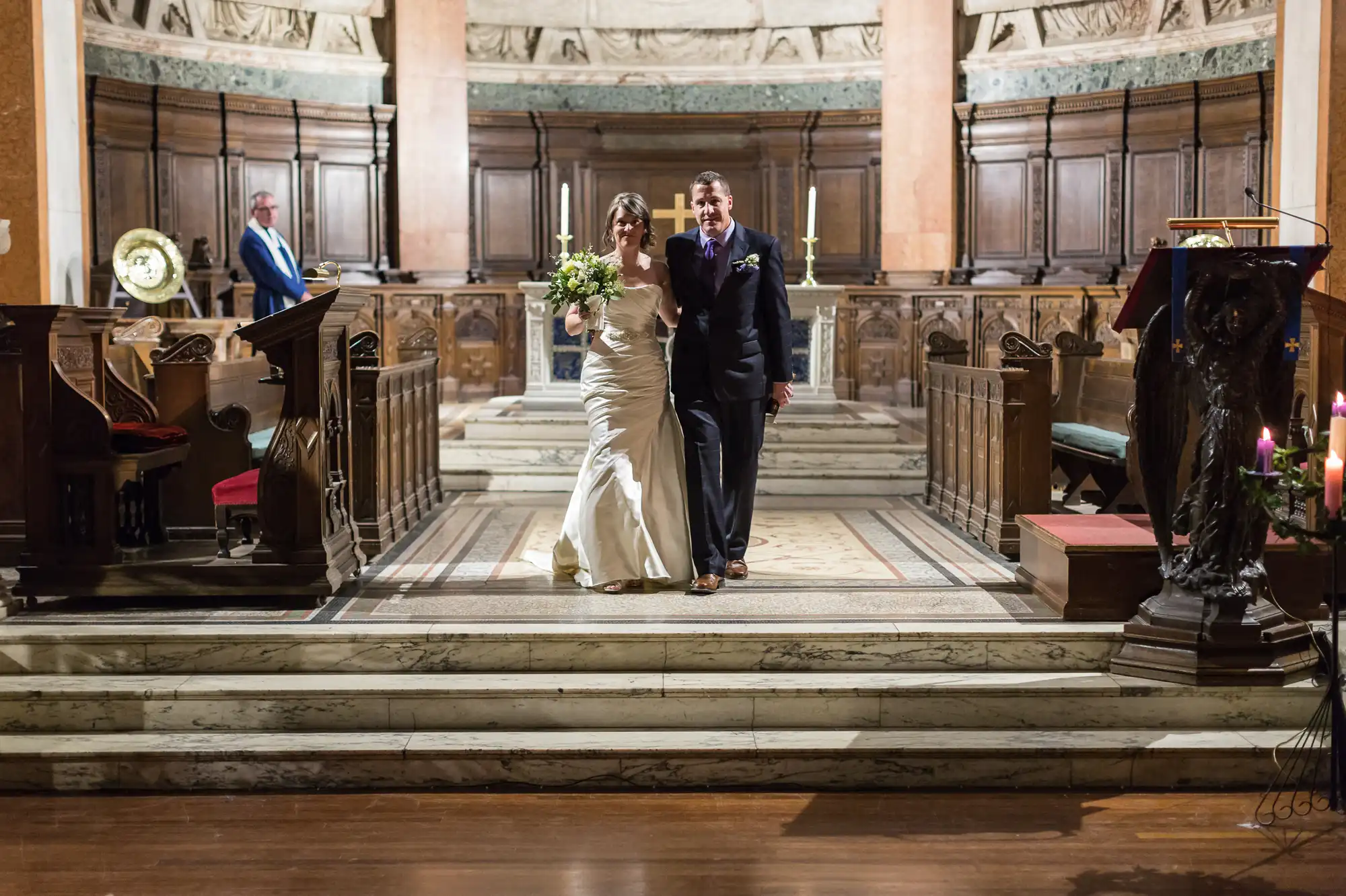 A bride and groom smiling and walking down the aisle of an ornate church, the bride holding a bouquet, with lit candles and guests around.