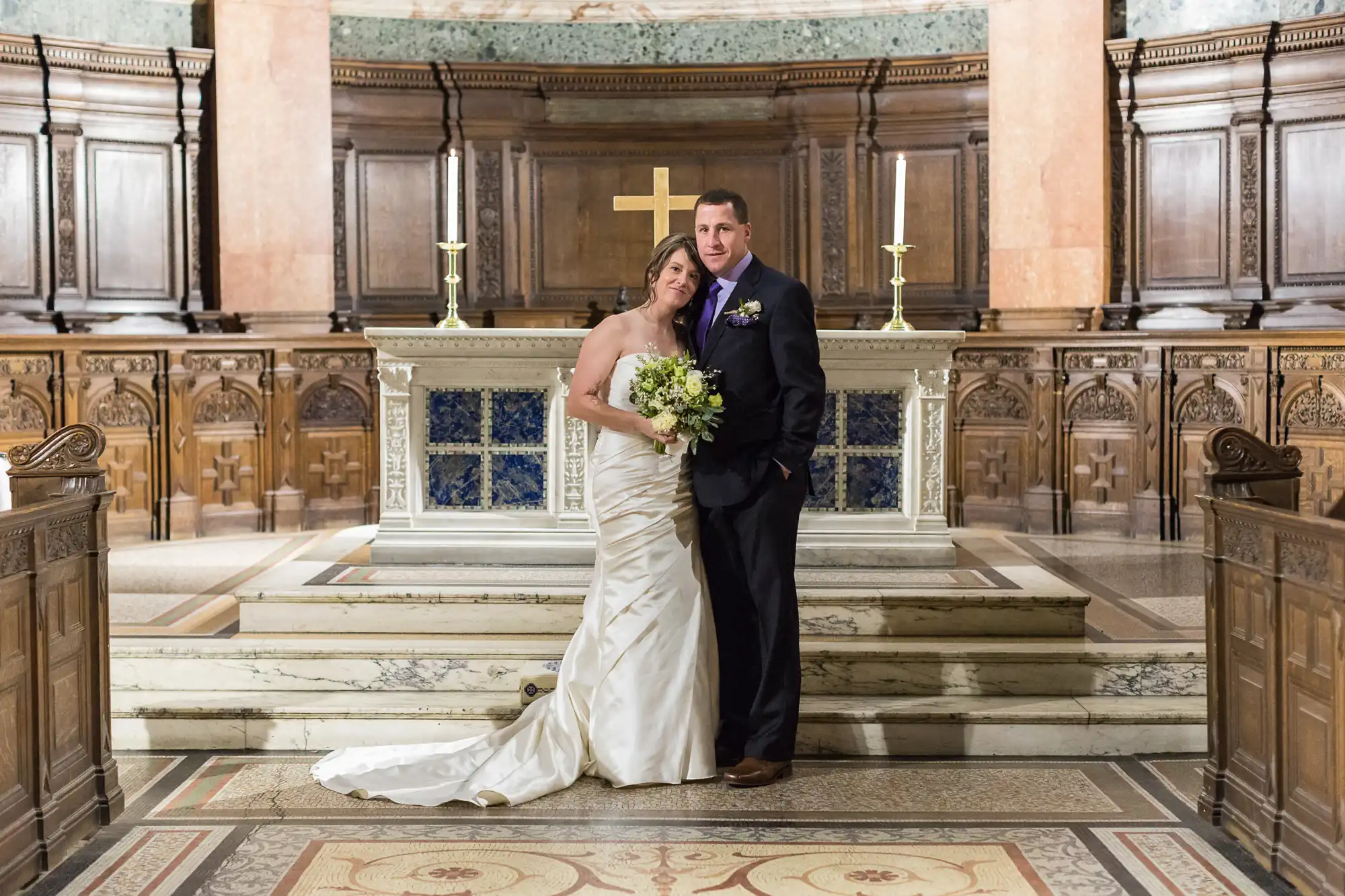 A bride and groom stand together in a grand church, smiling, with the groom embracing the bride from behind.