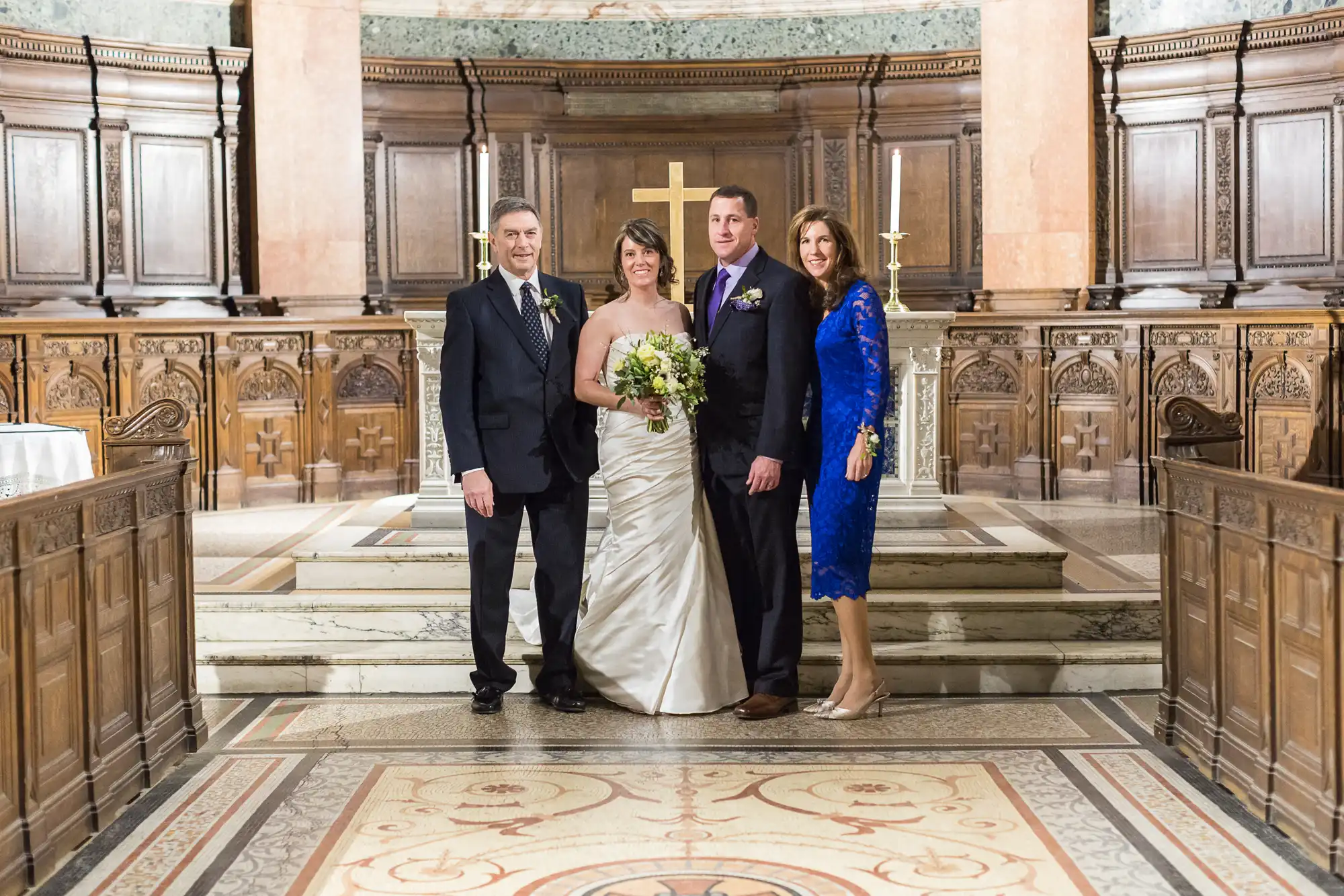 Two couples stand in a church, smiling, with a bride in a white dress holding a bouquet, flanked by two men in suits and a woman in a blue dress.