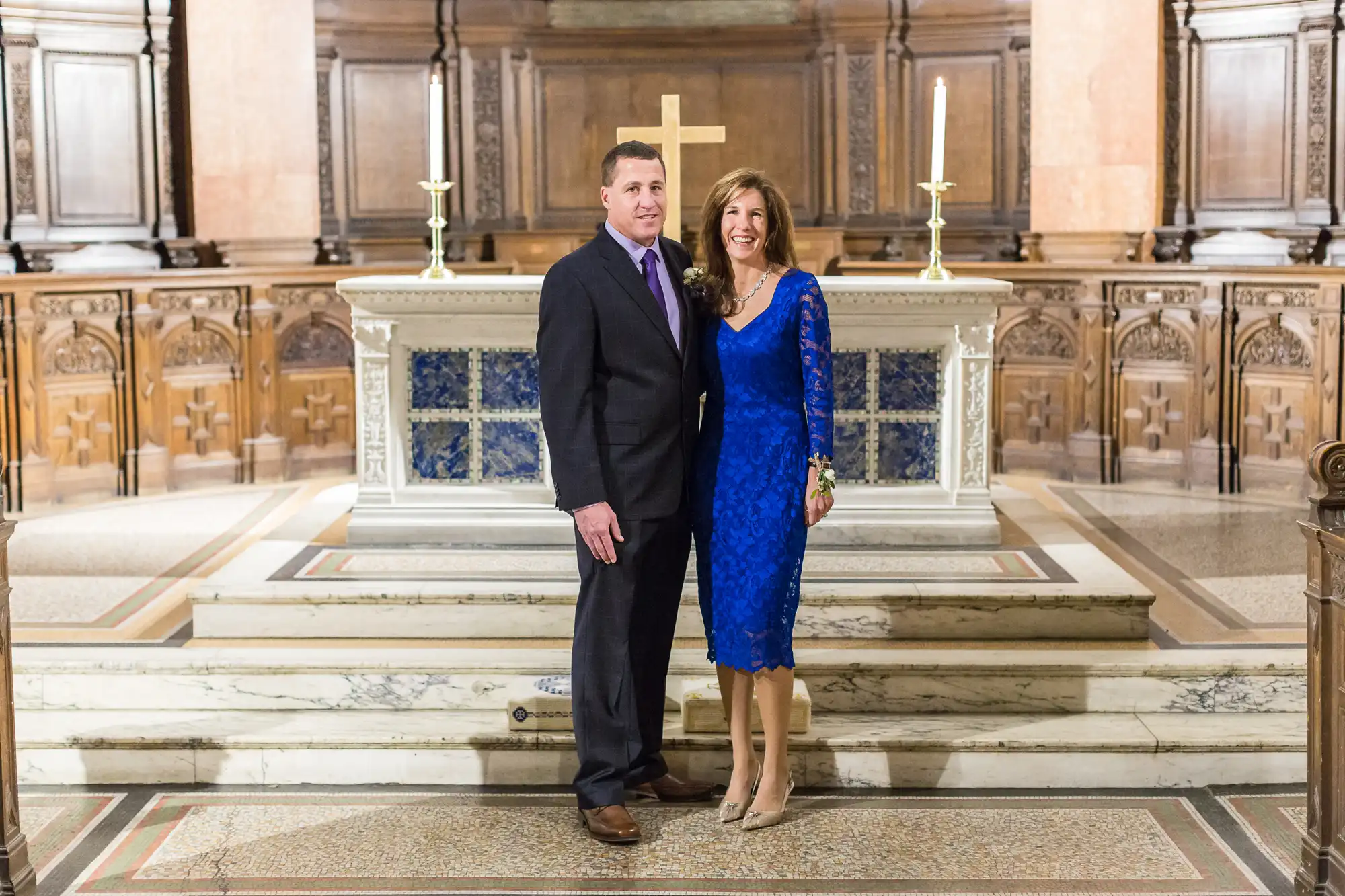 Man and woman in formal attire smiling in a church, standing in front of an altar with lit candles.