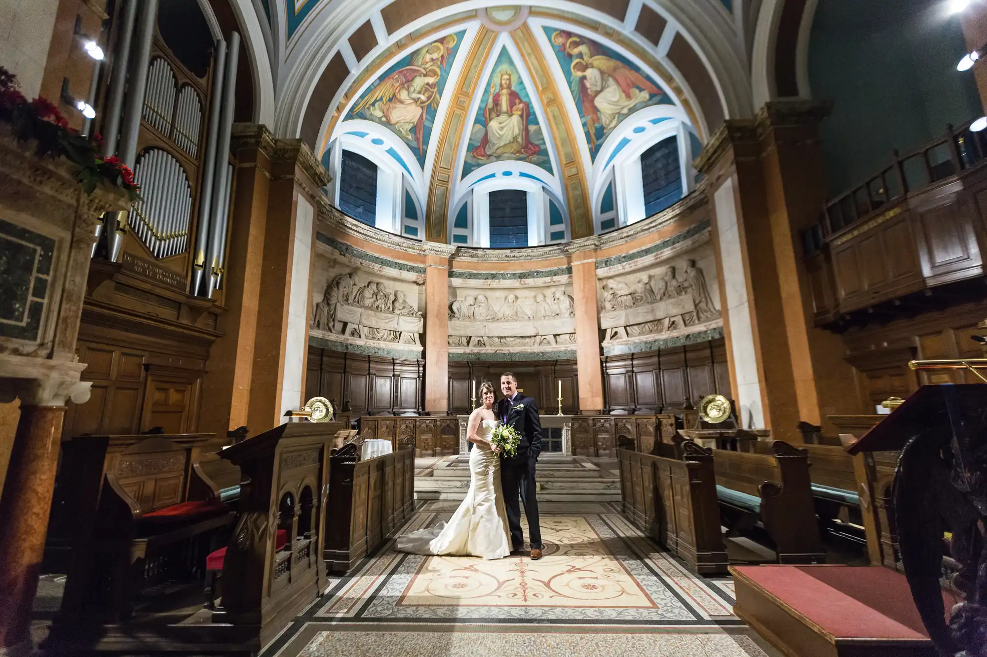 A bride and groom standing in the aisle of an ornate church with painted domed ceilings, colorful murals, and wooden pews.