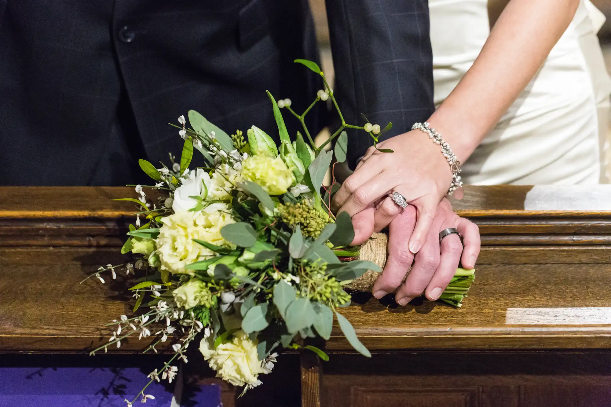 A newlywed couple gently holds hands over a church pew, showcasing their wedding rings and a bouquet of white and green flowers.