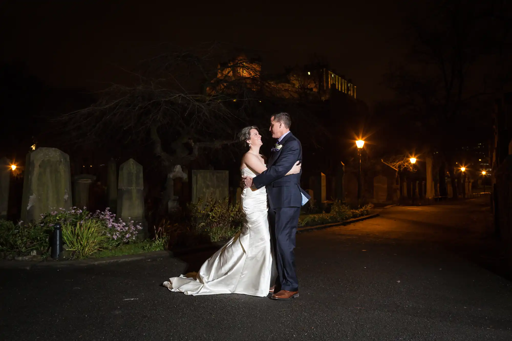 A bride and groom embrace at night in a lit cemetery, with headstones and illuminated paths surrounding them.