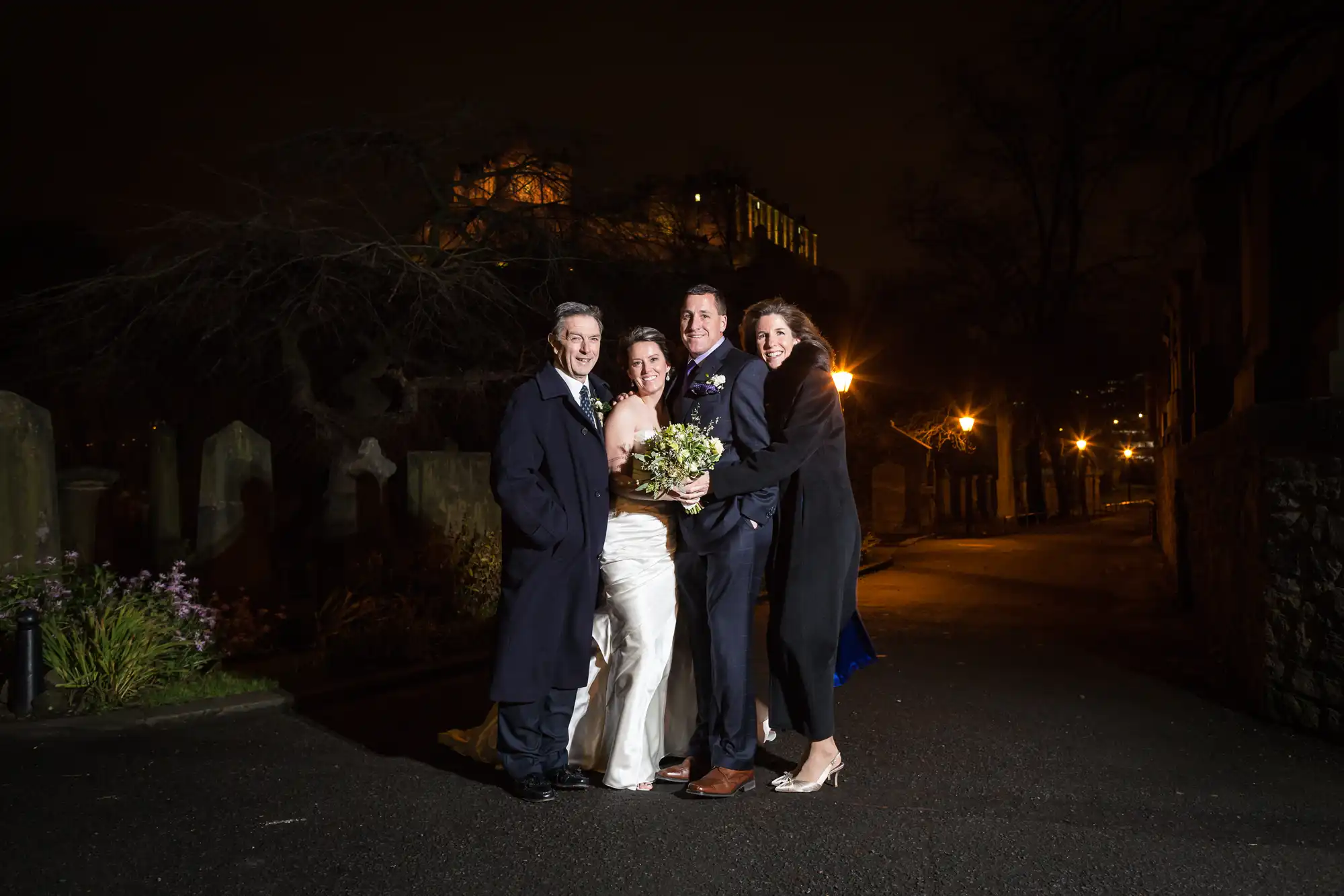 Four people dressed in formal attire posing for a photo at night on a street, with a dimly lit building and trees in the background.