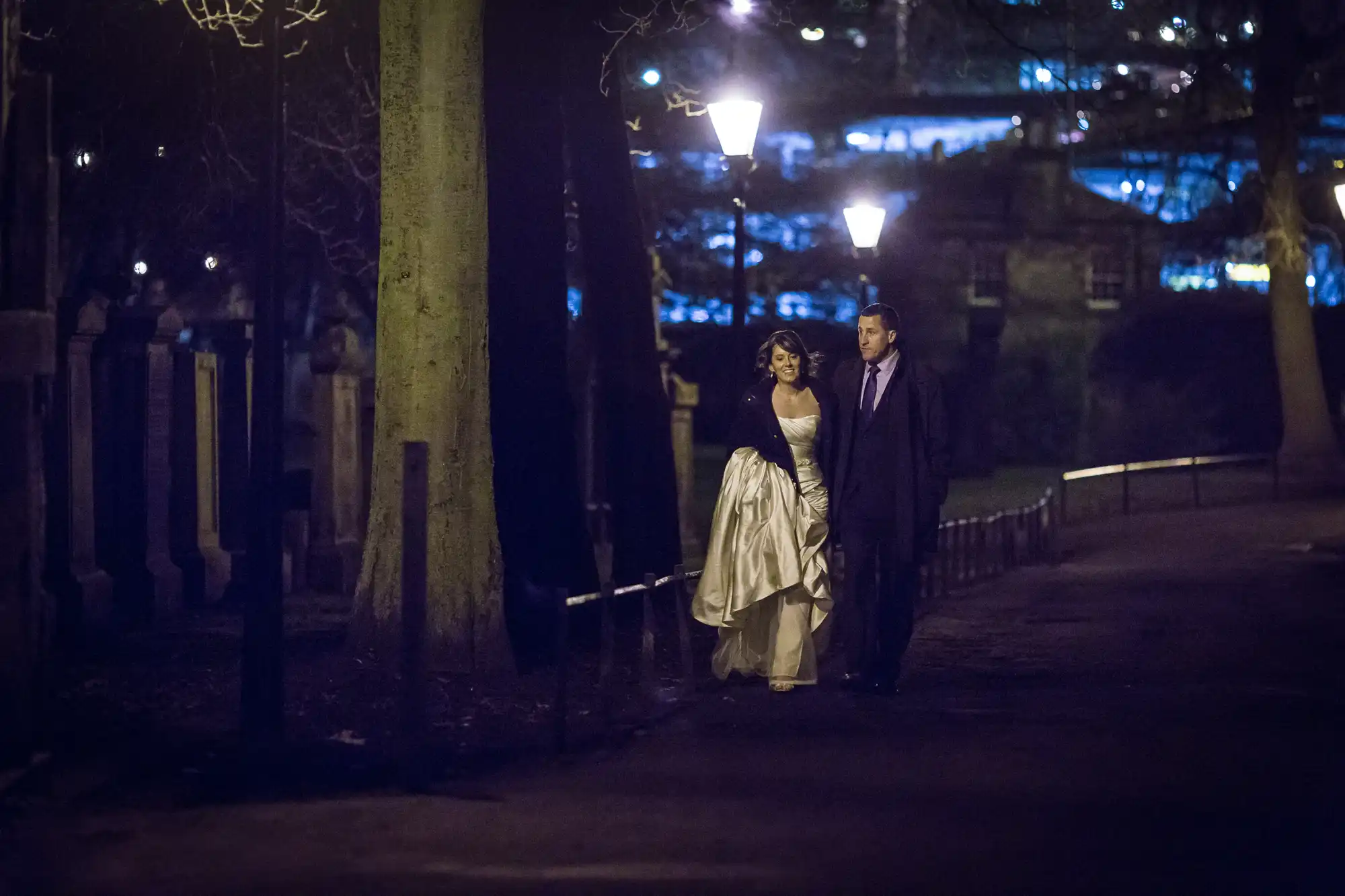 A couple in formal attire walking at night along a lamp-lit park path, with dim city lights visible in the background.