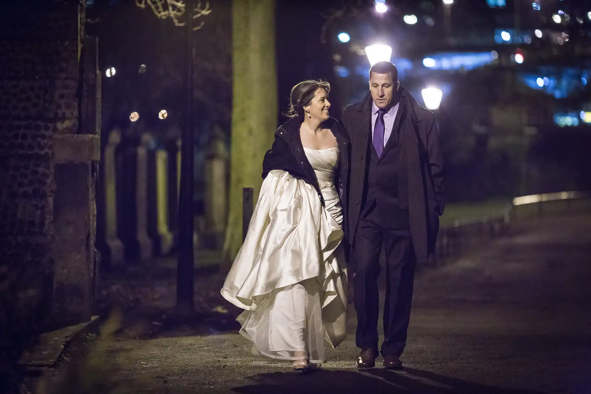 A bride in a white dress and a groom in a suit walk hand in hand along a dimly lit path at night, smiling and talking.