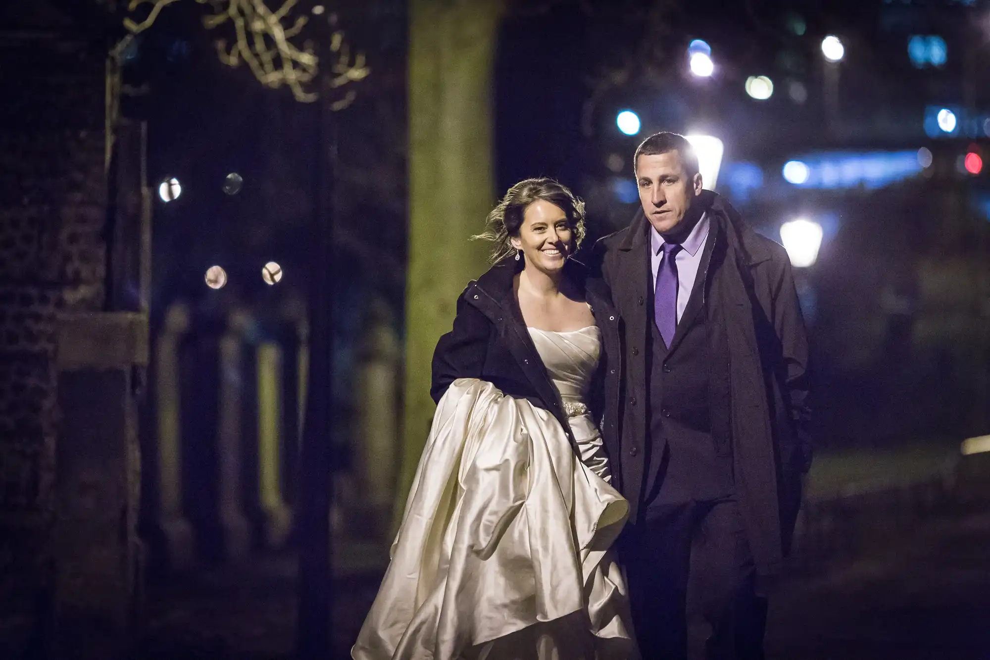 A bride in a white dress and a man in a suit and overcoat walk together at night, smiling, with streetlights in the background.