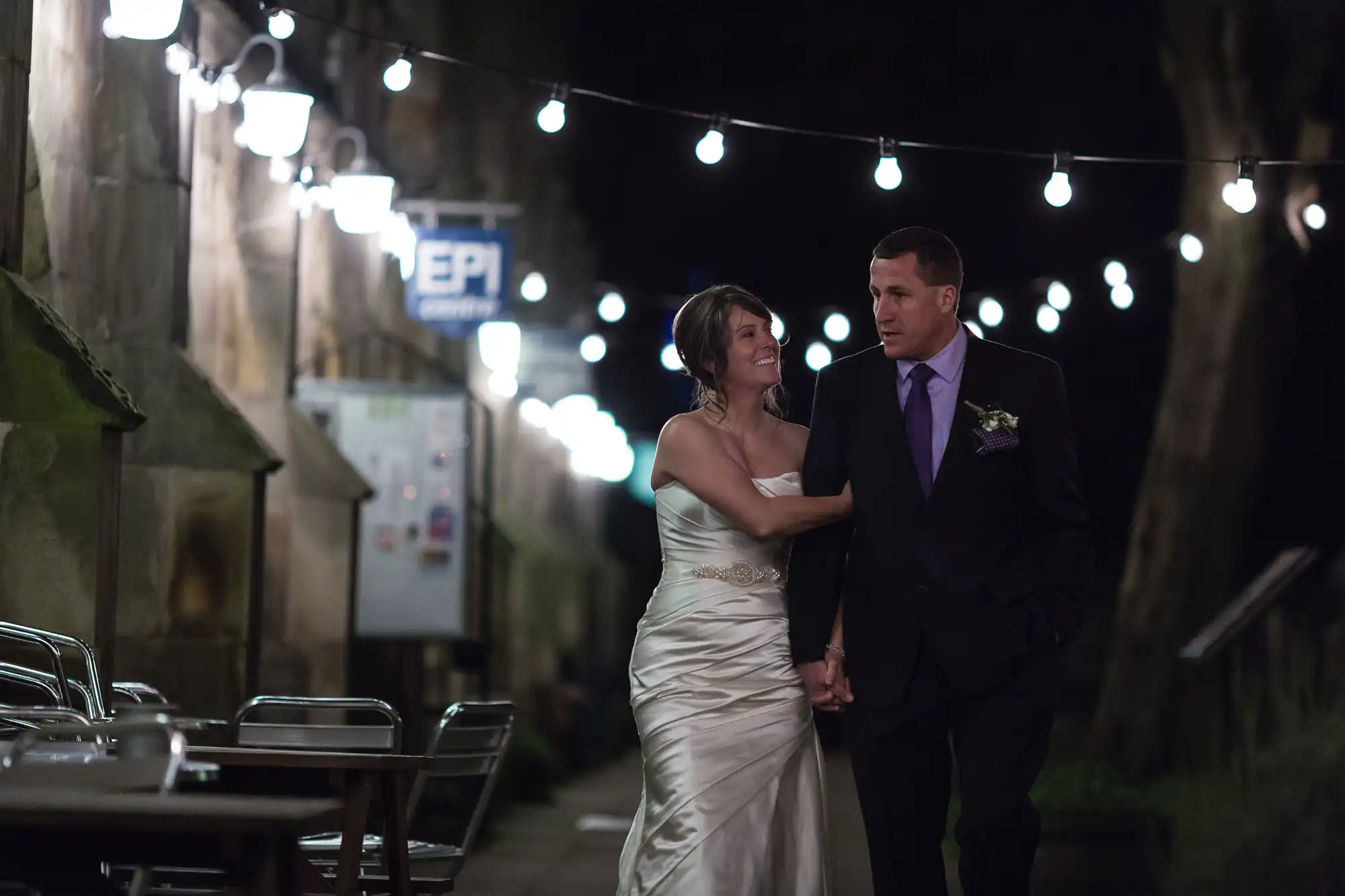 A bride and groom walk hand in hand at night along a warmly lit, narrow street adorned with string lights.