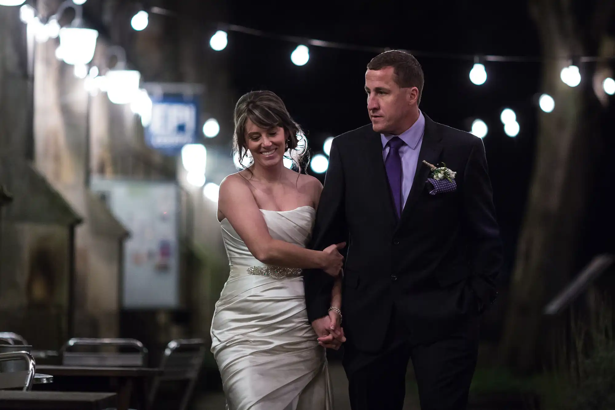 A newlywed couple walking under string lights at night, the bride smiling and linking arms with the groom who wears a purple tie.