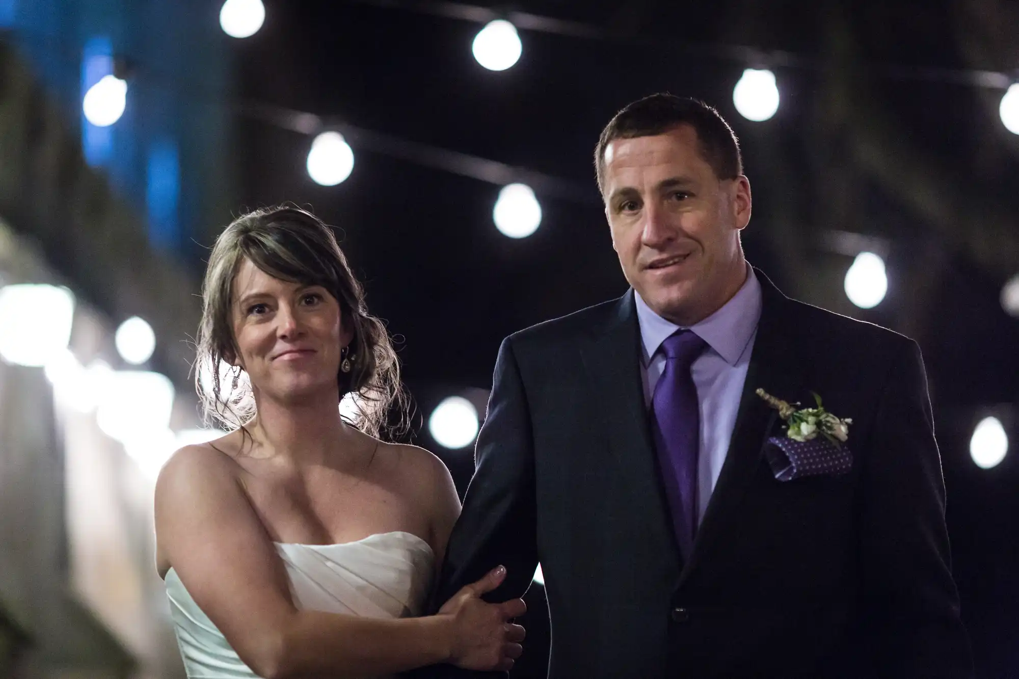 A bride in a white dress and a groom in a dark suit walk arm-in-arm at night under string lights.