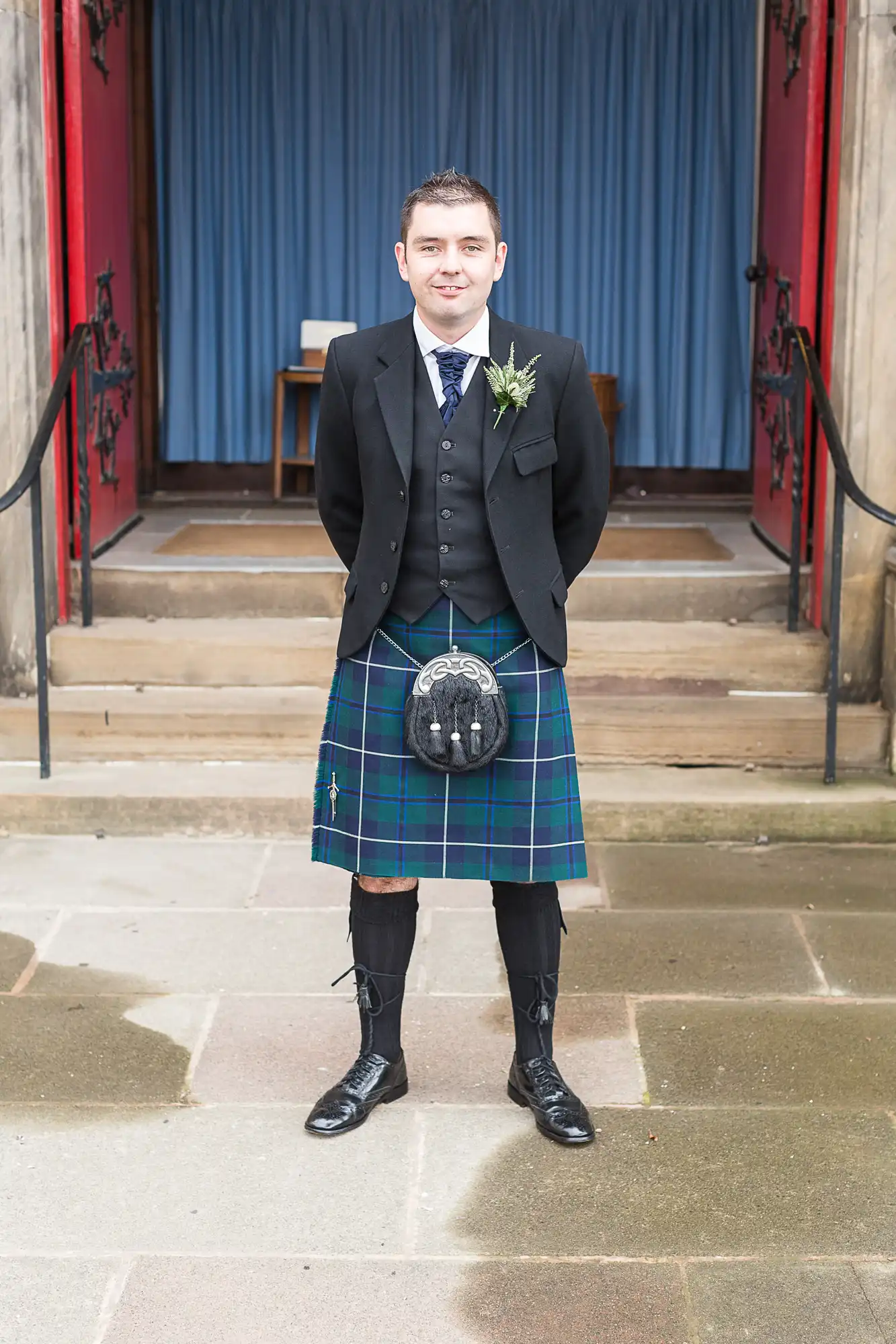 Man in traditional scottish kilt and jacket standing confidently in front of a church entrance with blue doors.