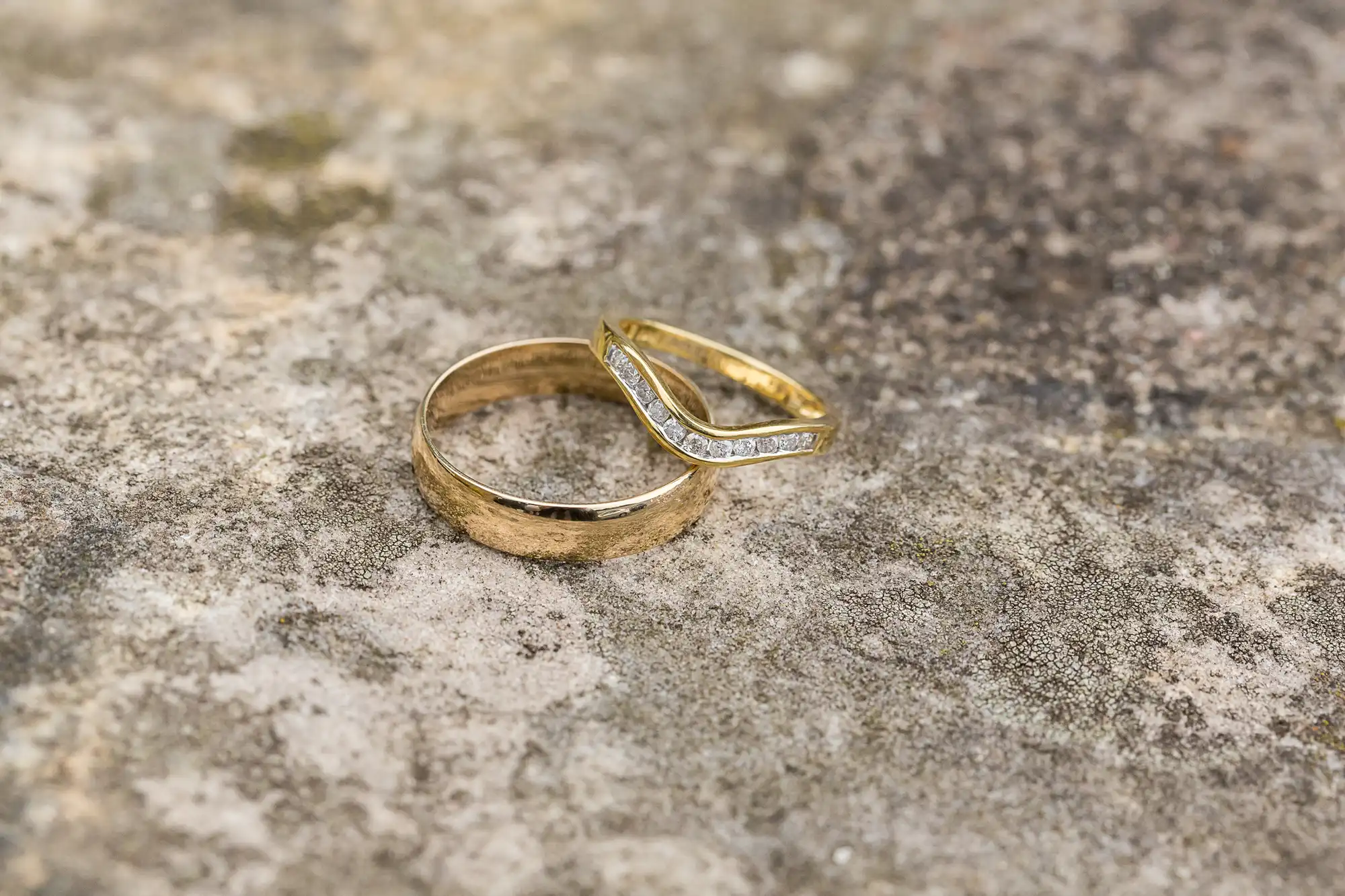 Two wedding rings, one plain gold and one gold with diamonds, resting on a textured stone surface.