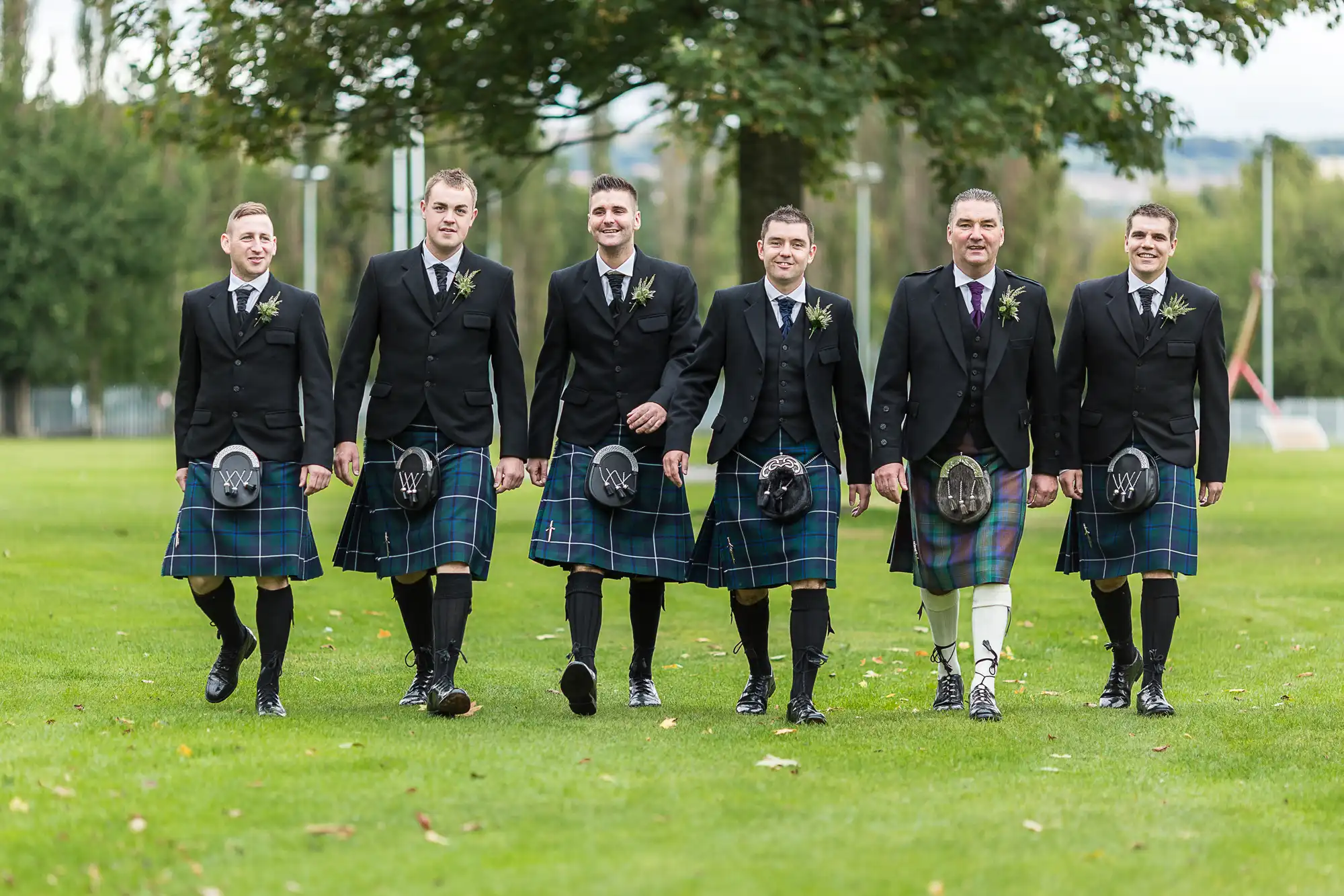 Six men in traditional scottish kilts and jackets walking side by side on a grassy field, smiling.