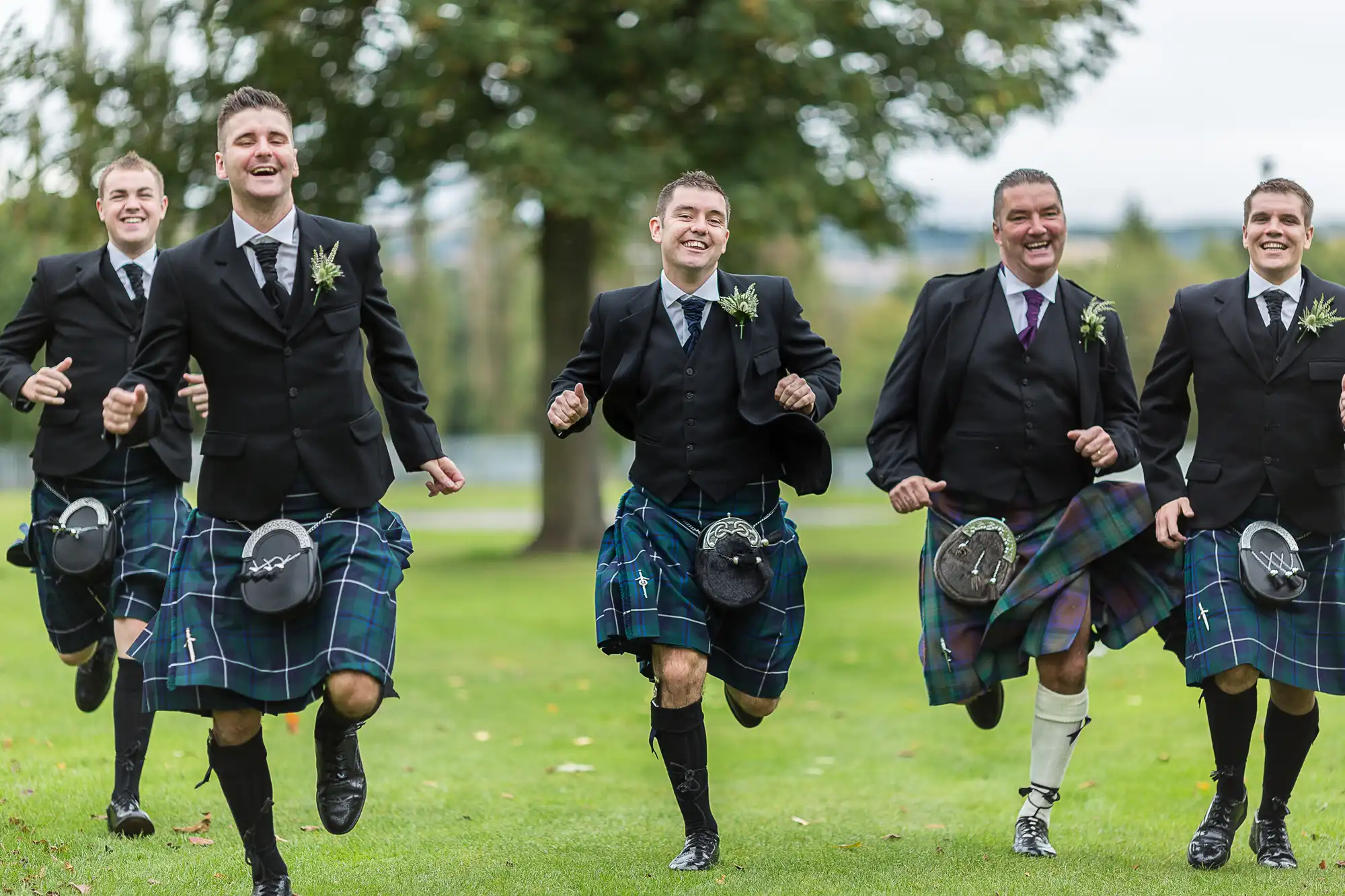 Four men in scottish kilts and jackets joyfully running across a grassy field, smiling broadly.