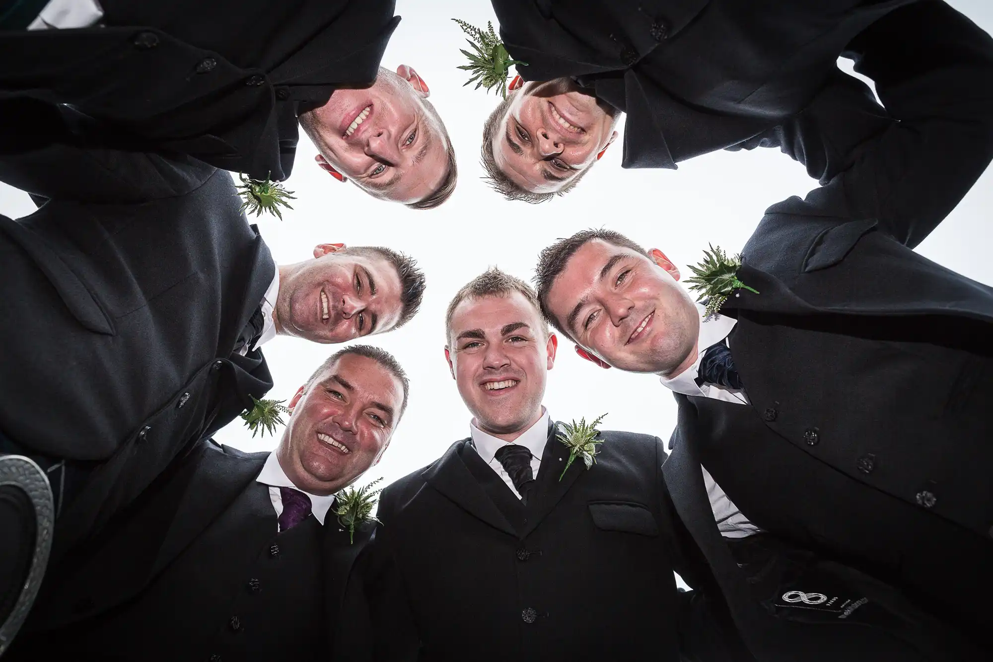 Five men in formal attire with boutonnieres, smiling down at the camera in a circle, photographed from a low angle.