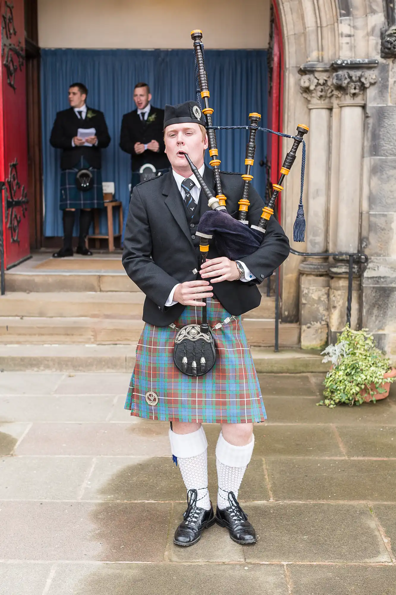 A man in traditional scottish attire plays the bagpipes at an outdoor event, wearing a tartan kilt and black jacket.