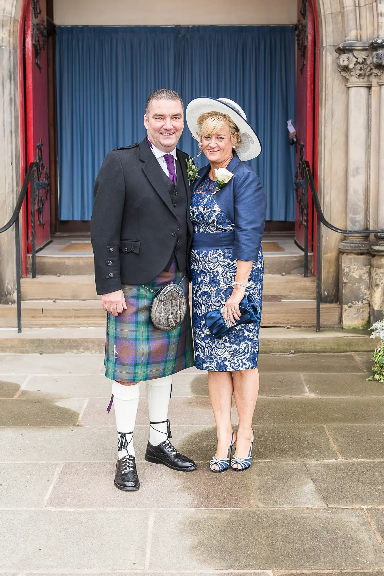 A man in a kilt and jacket with a woman in a blue dress and hat, both smiling, standing in front of a church entrance with blue doors.