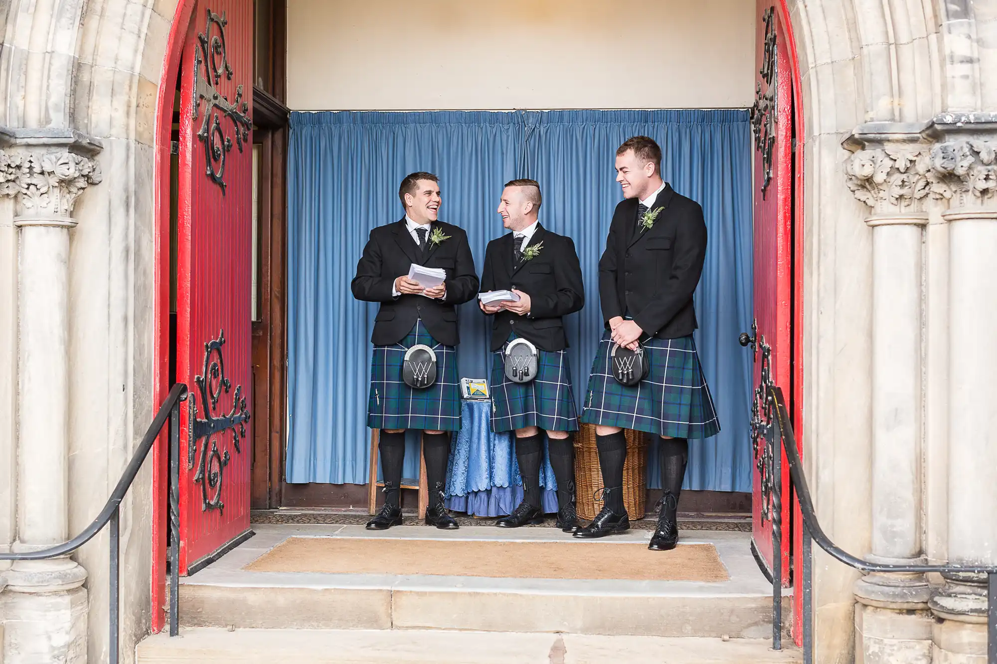 Three men in traditional scottish kilts and accessories stand laughing at a church doorway, with one holding a ceremonial booklet.