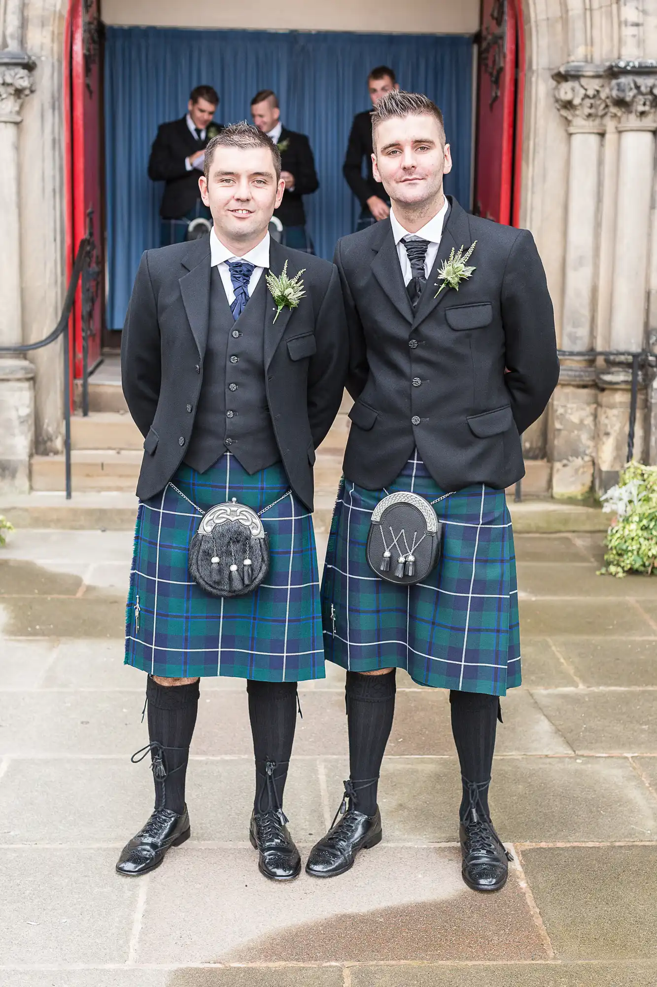 Two men in traditional scottish kilts and jackets, smiling and standing outside a building entrance.