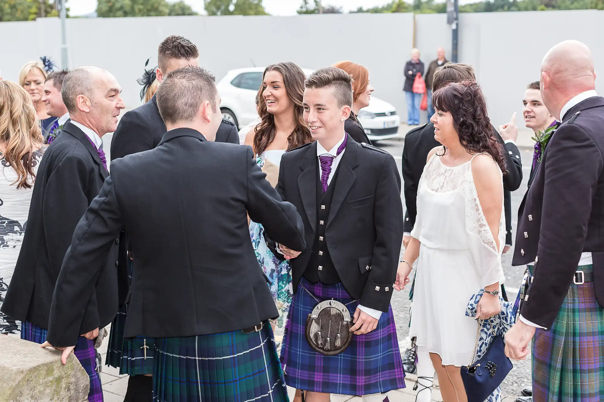 Group of people in formal attire, including kilts, gathered in a parking lot, greeting each other with handshakes and smiles.