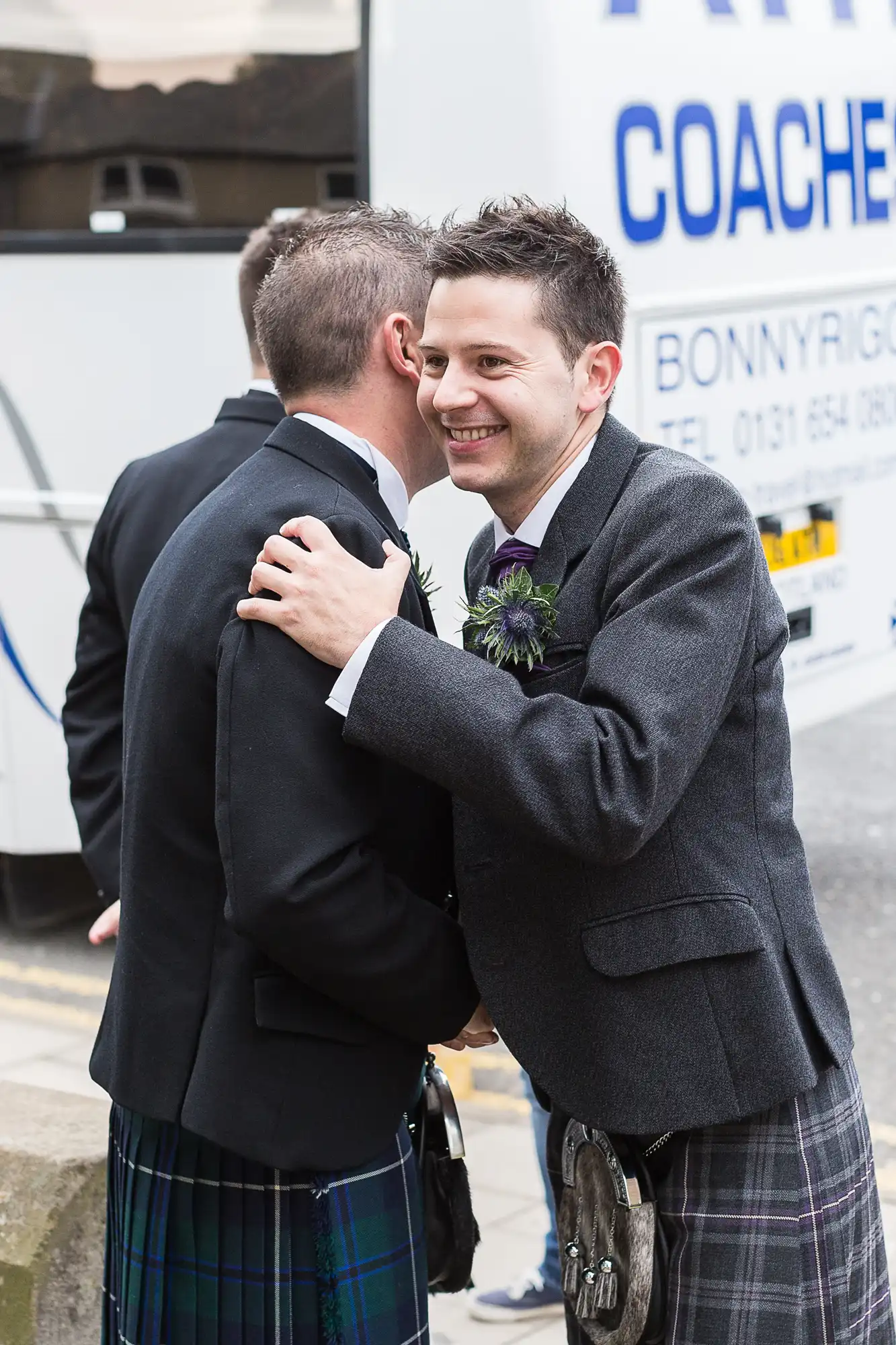 Two men in kilts embracing and smiling at a festive event, one adjusting the other's boutonniere.
