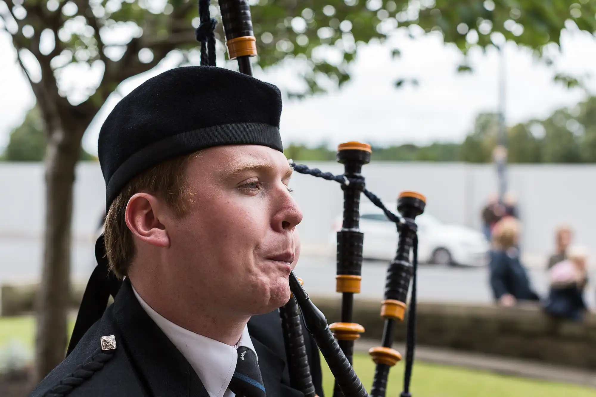 A man in a traditional scottish outfit playing the bagpipes outdoors, with a focused expression and people sitting in the background.