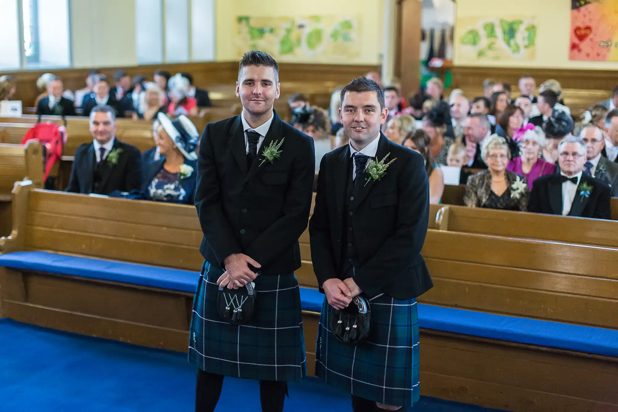 Two men in traditional kilts and jackets standing in a church aisle, with wedding guests seated in the background.