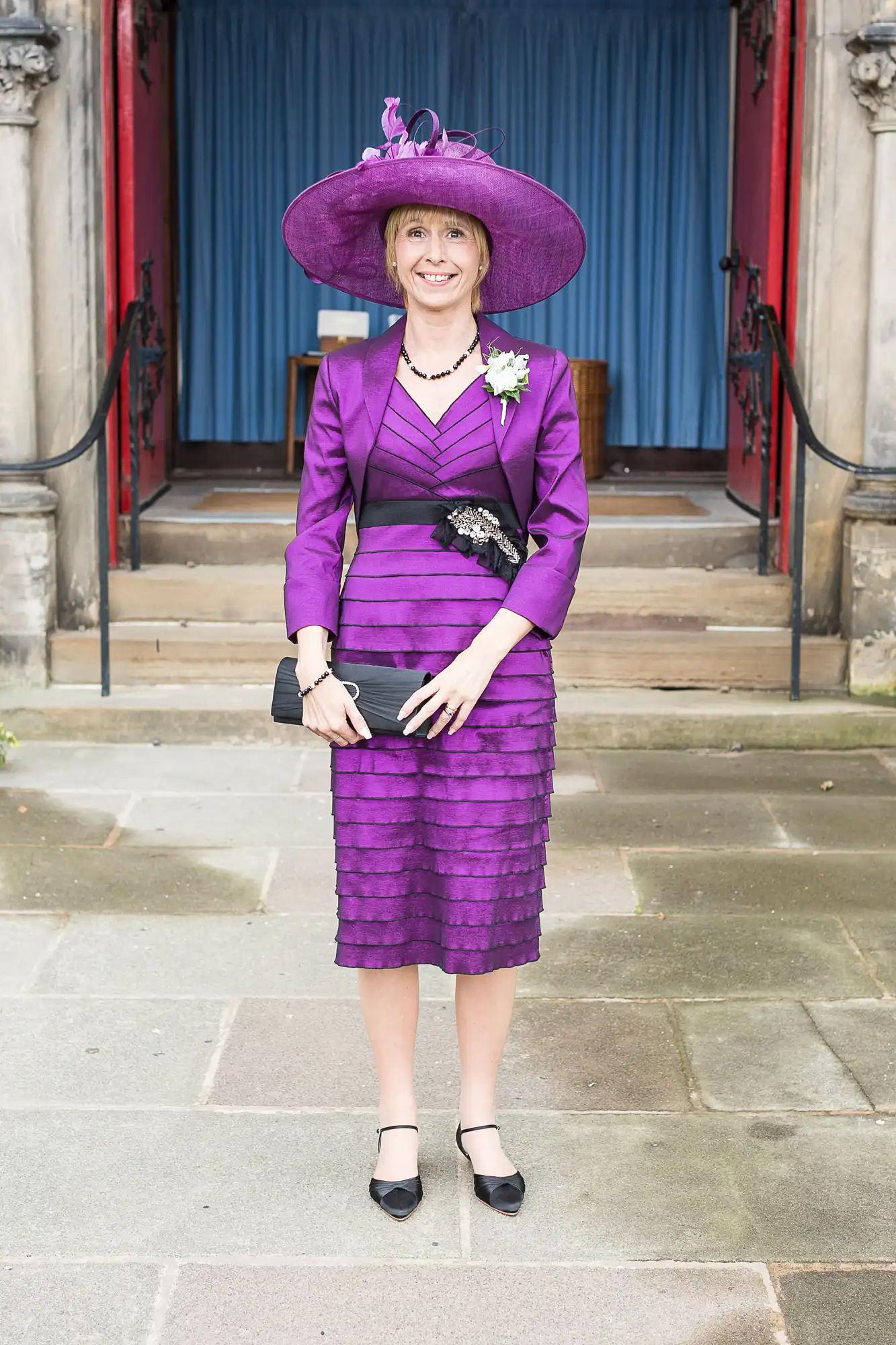 A woman in a purple dress and hat stands smiling in front of a building with red doors.