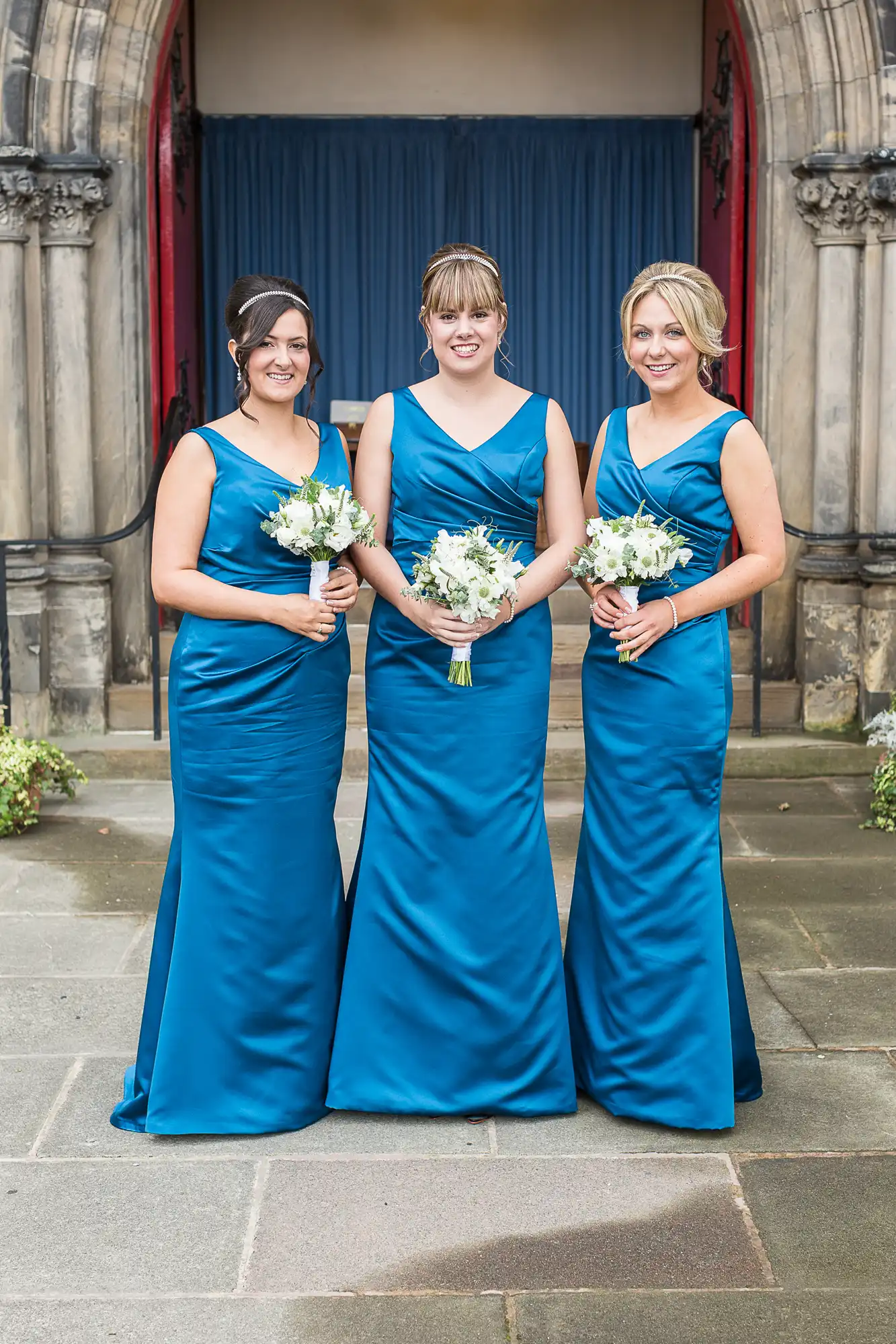Three women in matching blue dresses holding bouquets, posing in front of an old church entrance.