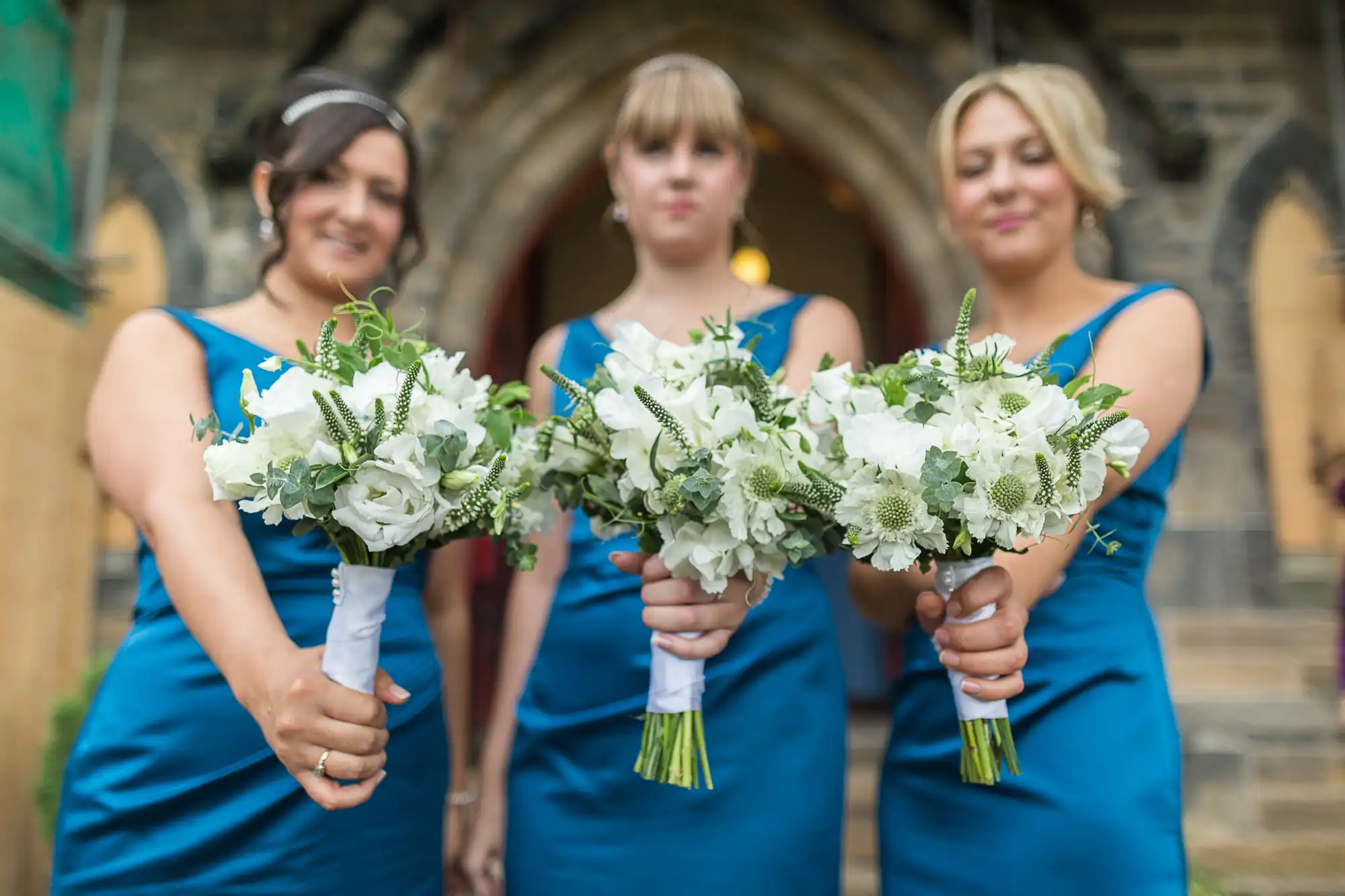 Three bridesmaids in blue dresses holding white floral bouquets in front of a church entrance.