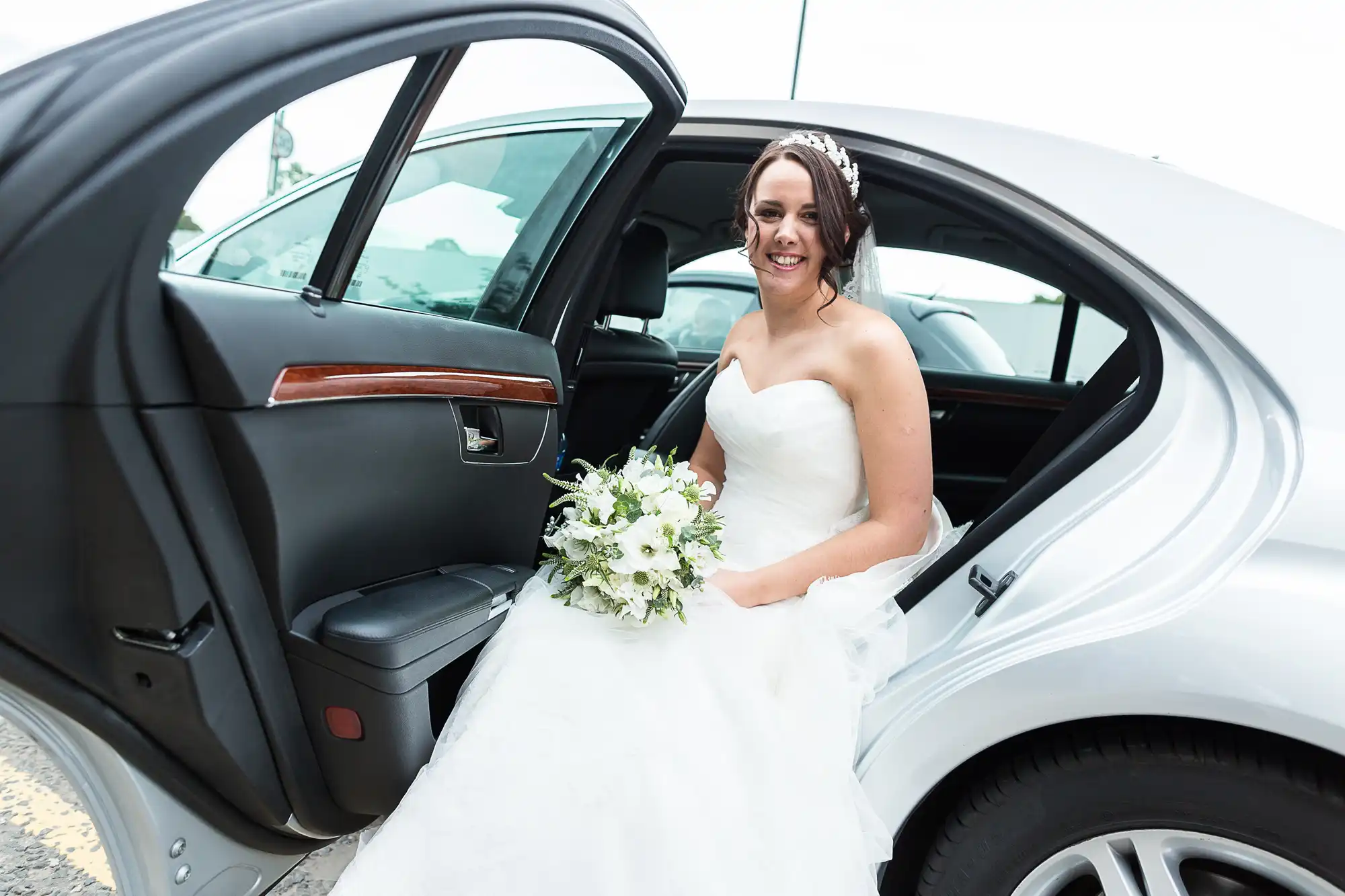 A bride in a white gown sitting inside a car, holding a bouquet, smiling at the camera through the open door.