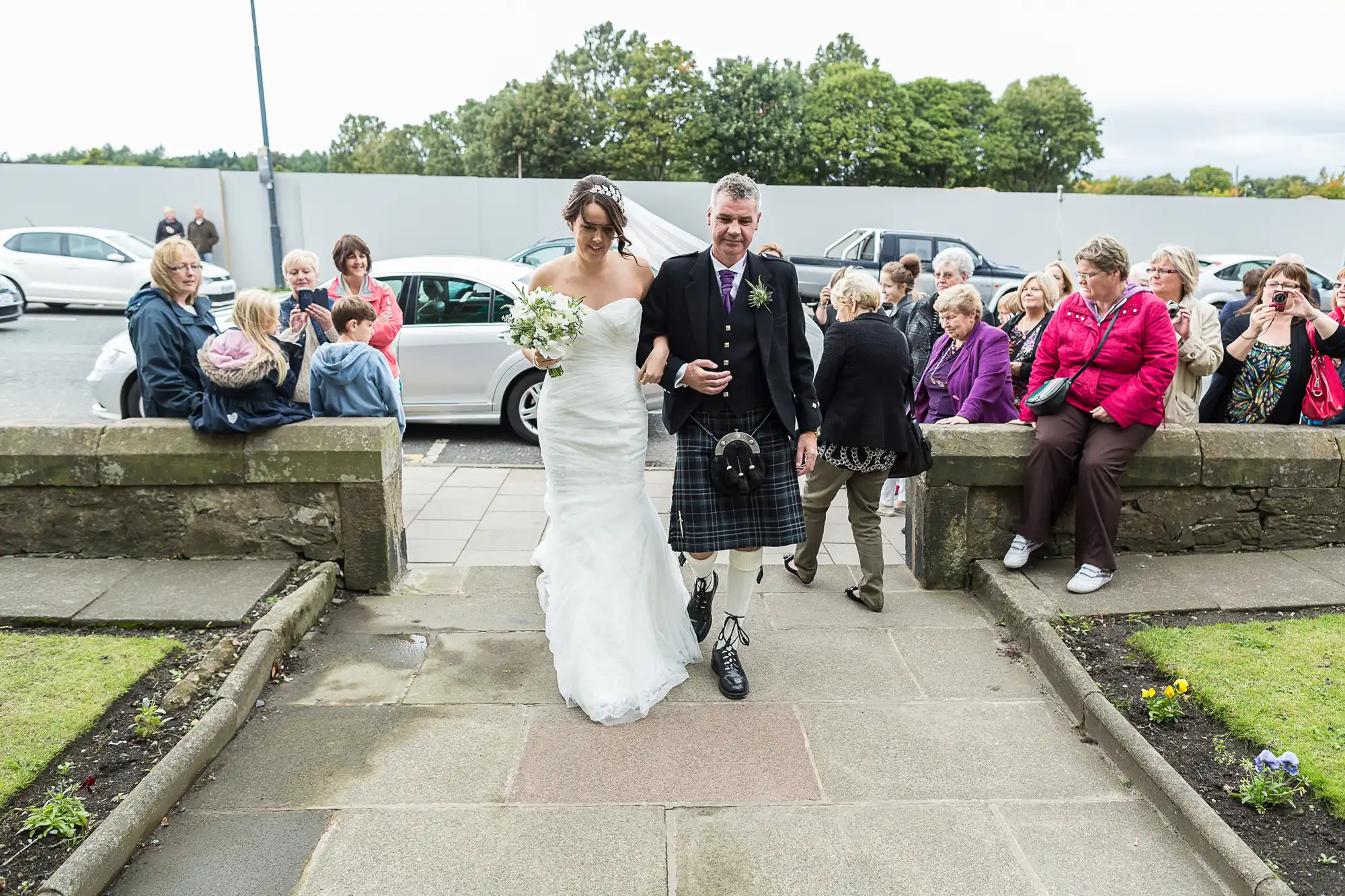 A bride in a white dress and a groom in a kilt walk hand in hand, surrounded by smiling onlookers, in an outdoor setting with parked cars in the background.