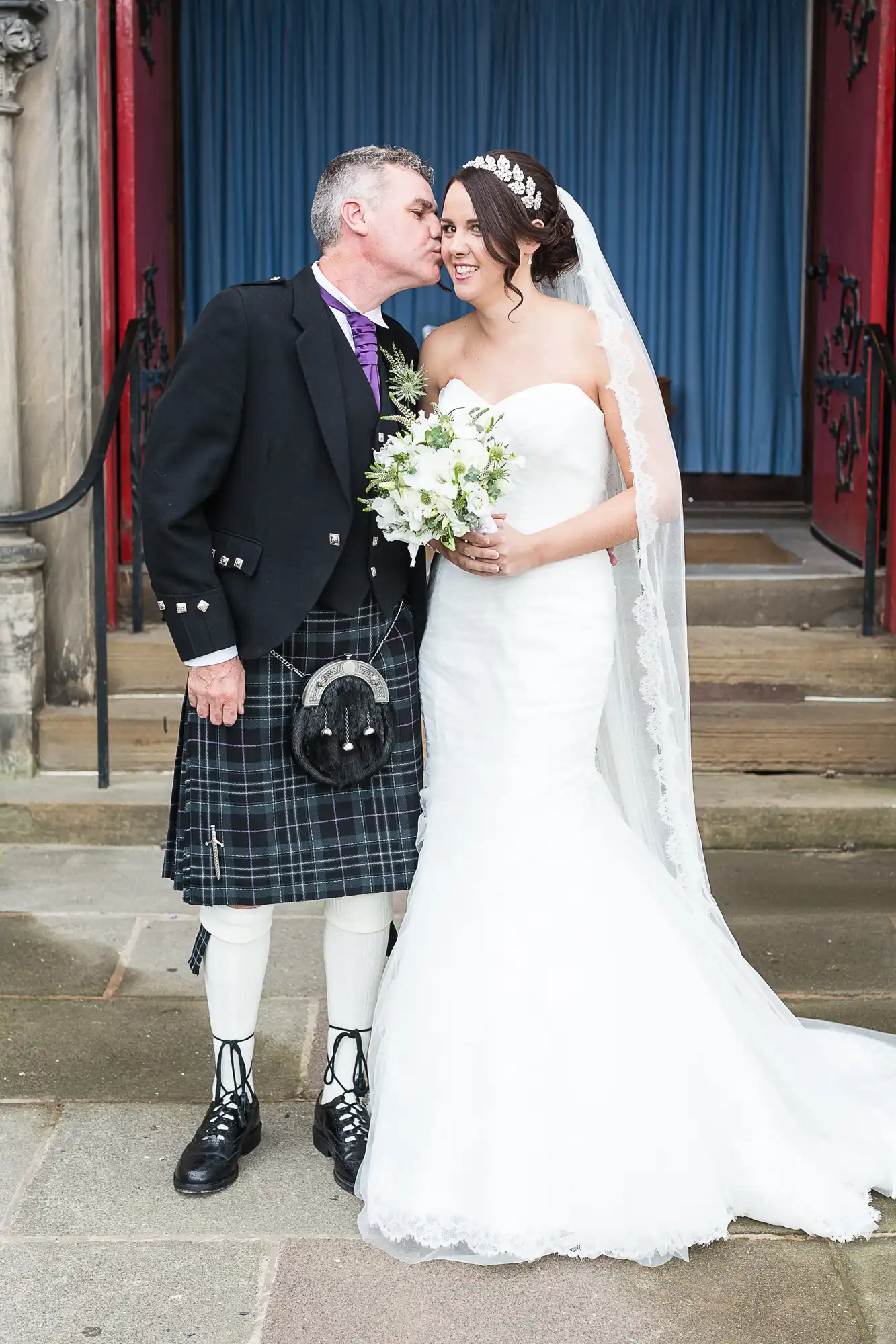 A groom in a kilt kisses the bride on the cheek outside a church; she is in a white gown and holding a bouquet.