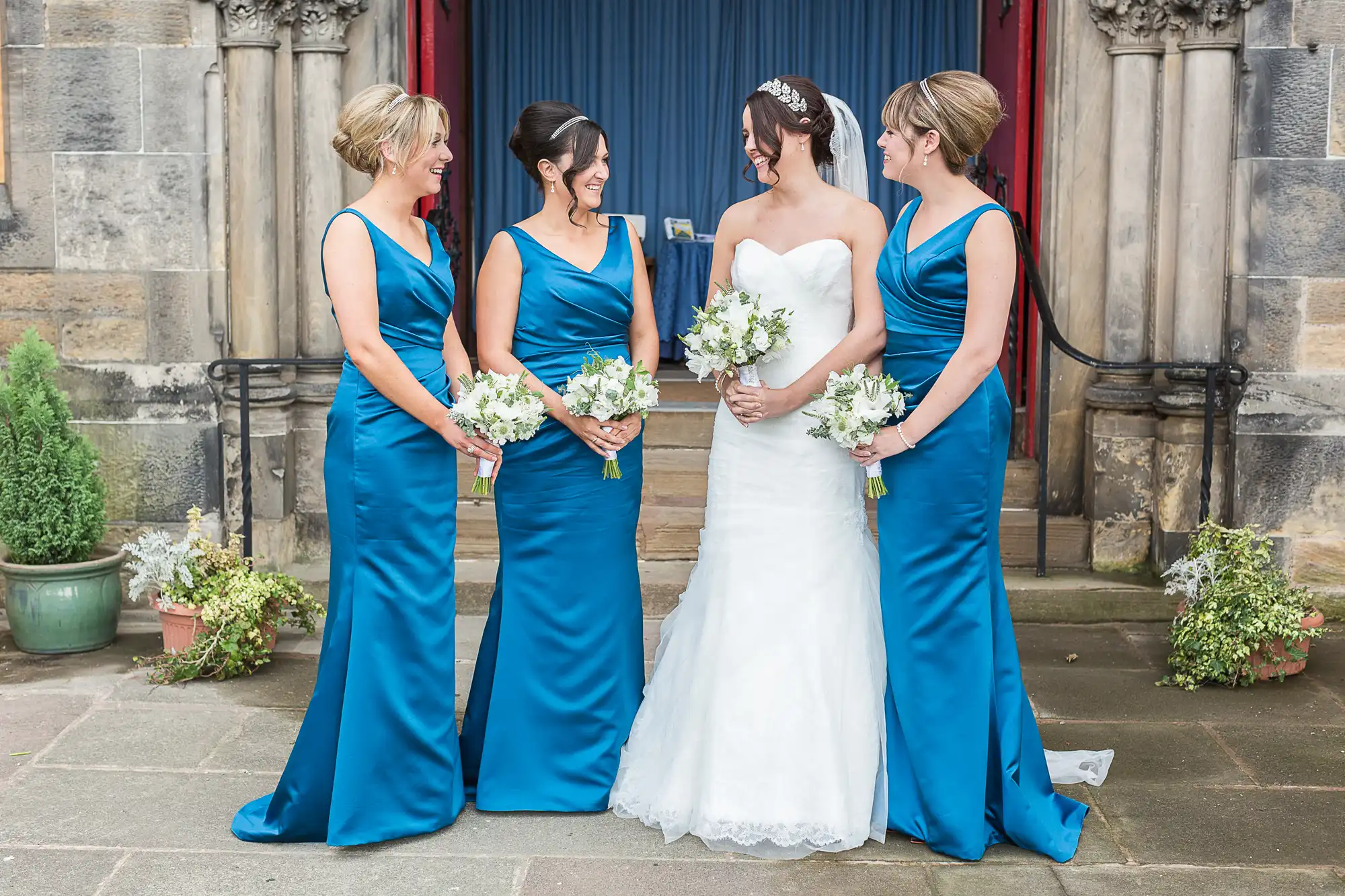 Four women in blue dresses, one in a white wedding gown, smiling and talking outside a church.