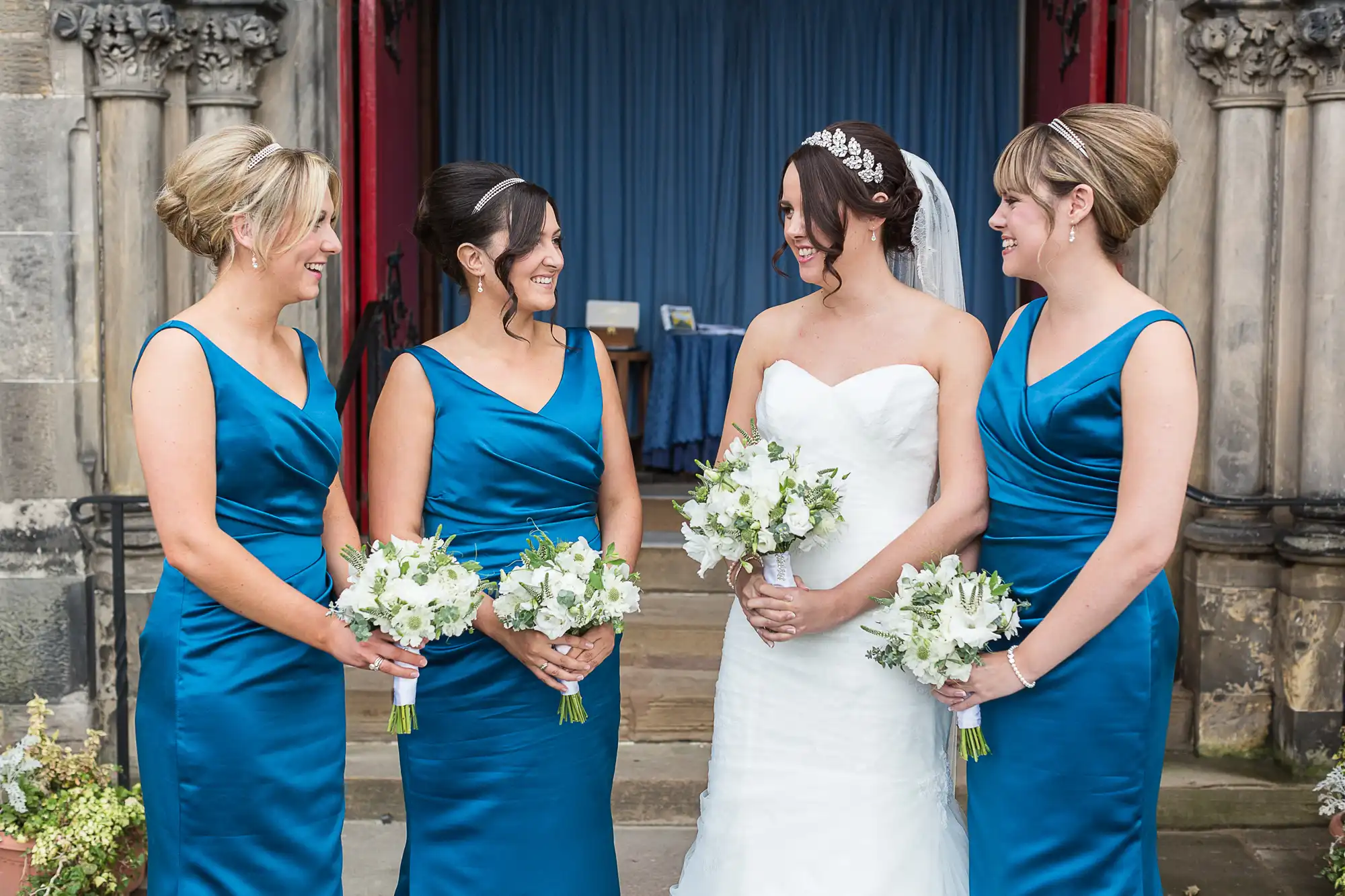 A bride in a white dress smiles at three bridesmaids in blue dresses, each holding a bouquet, standing outside a church.