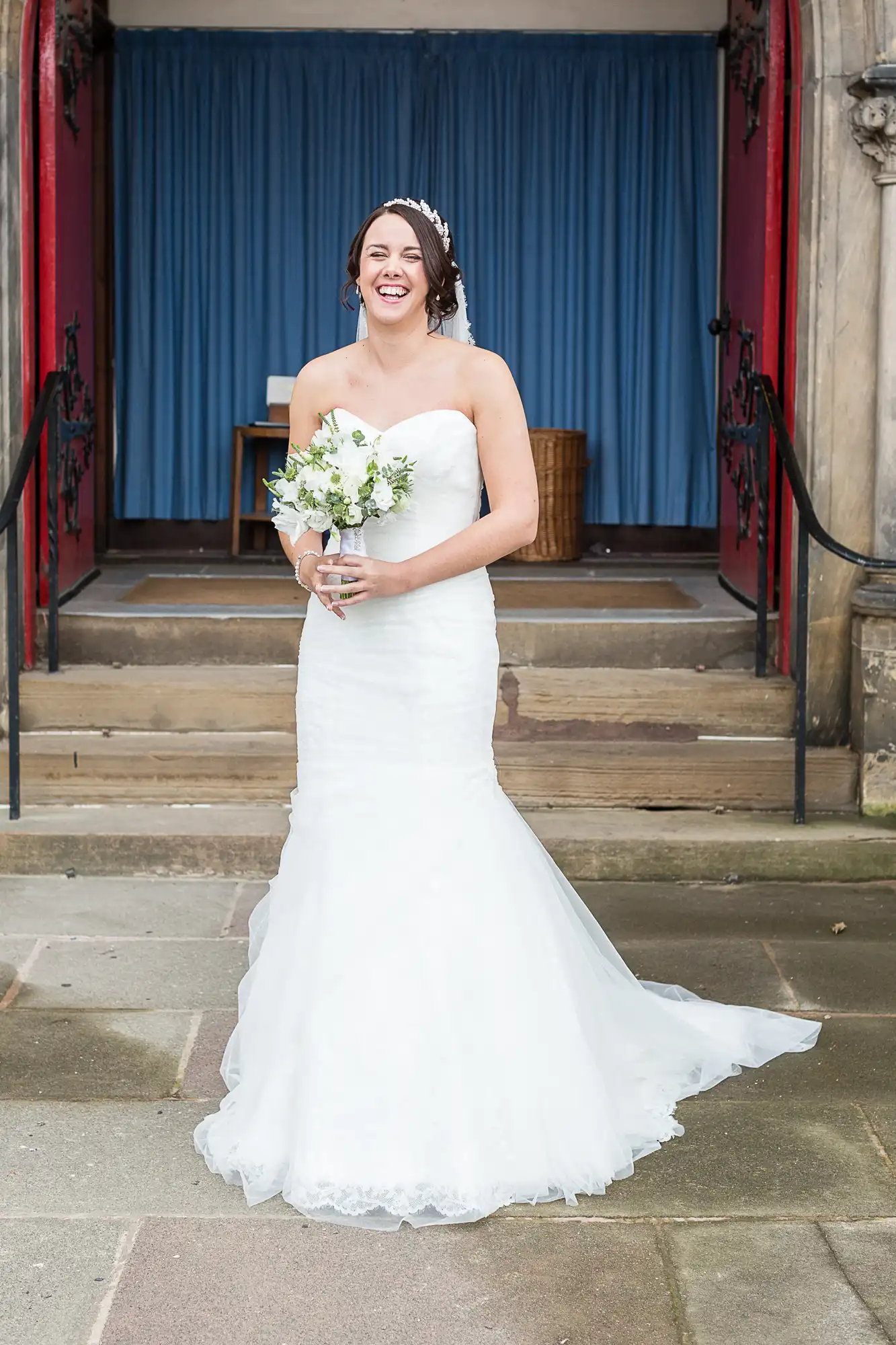 A bride in a strapless white gown laughs joyously, holding a bouquet, in front of a blue curtained doorway.