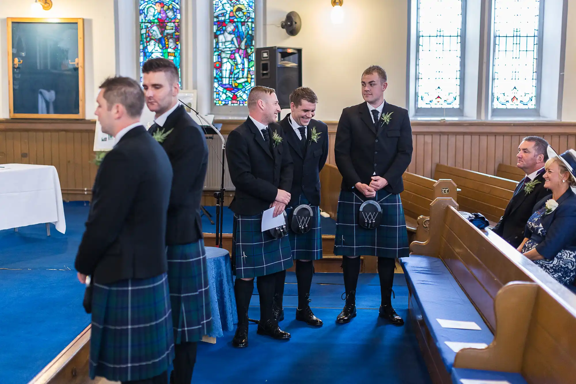 Men in traditional kilts standing inside a church during a wedding ceremony, some chatting and smiling.
